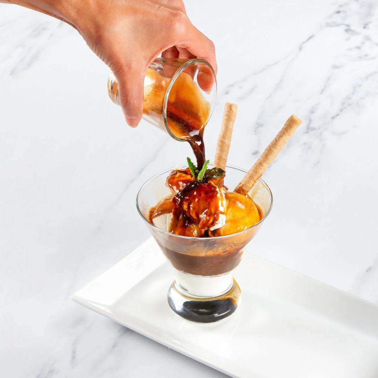 Pouring caramel sauce over ice cream sundaes with wafer sticks and mint leaves in a glass on a white plate and marble surface.