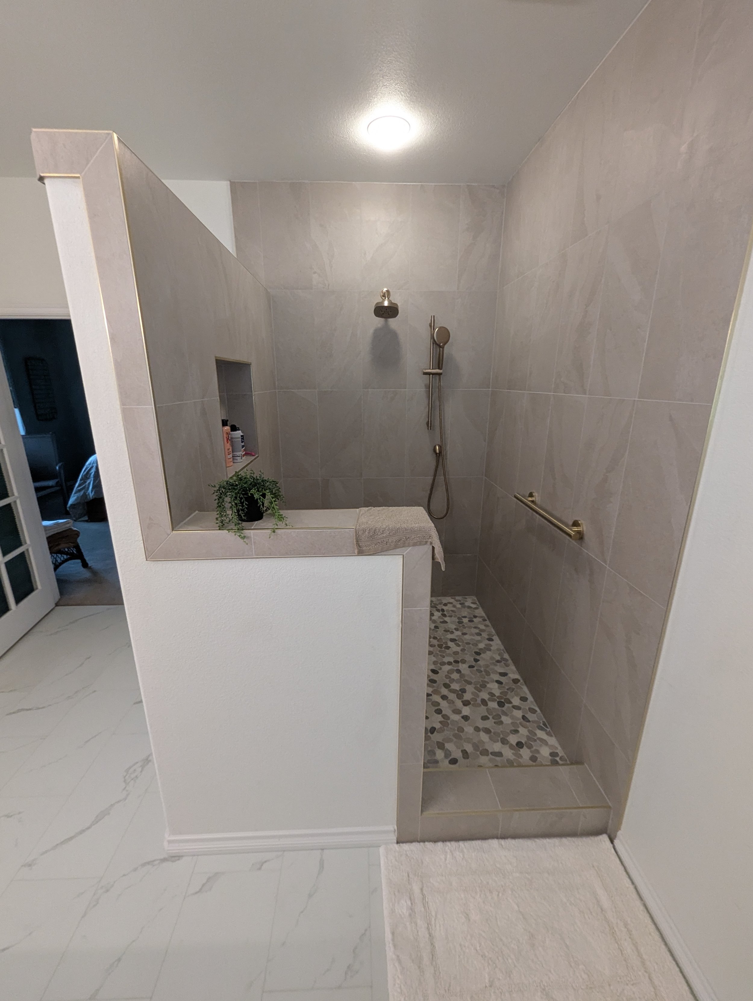 A bathroom shower area with beige tiled walls, a wall-mounted showerhead, a handheld shower hose, and a grab bar. There is a small built-in shelf with toiletries and a potted plant. The shower area has a pebble tile floor and is separated from the rest of the bathroom by a partial wall and a mirror.