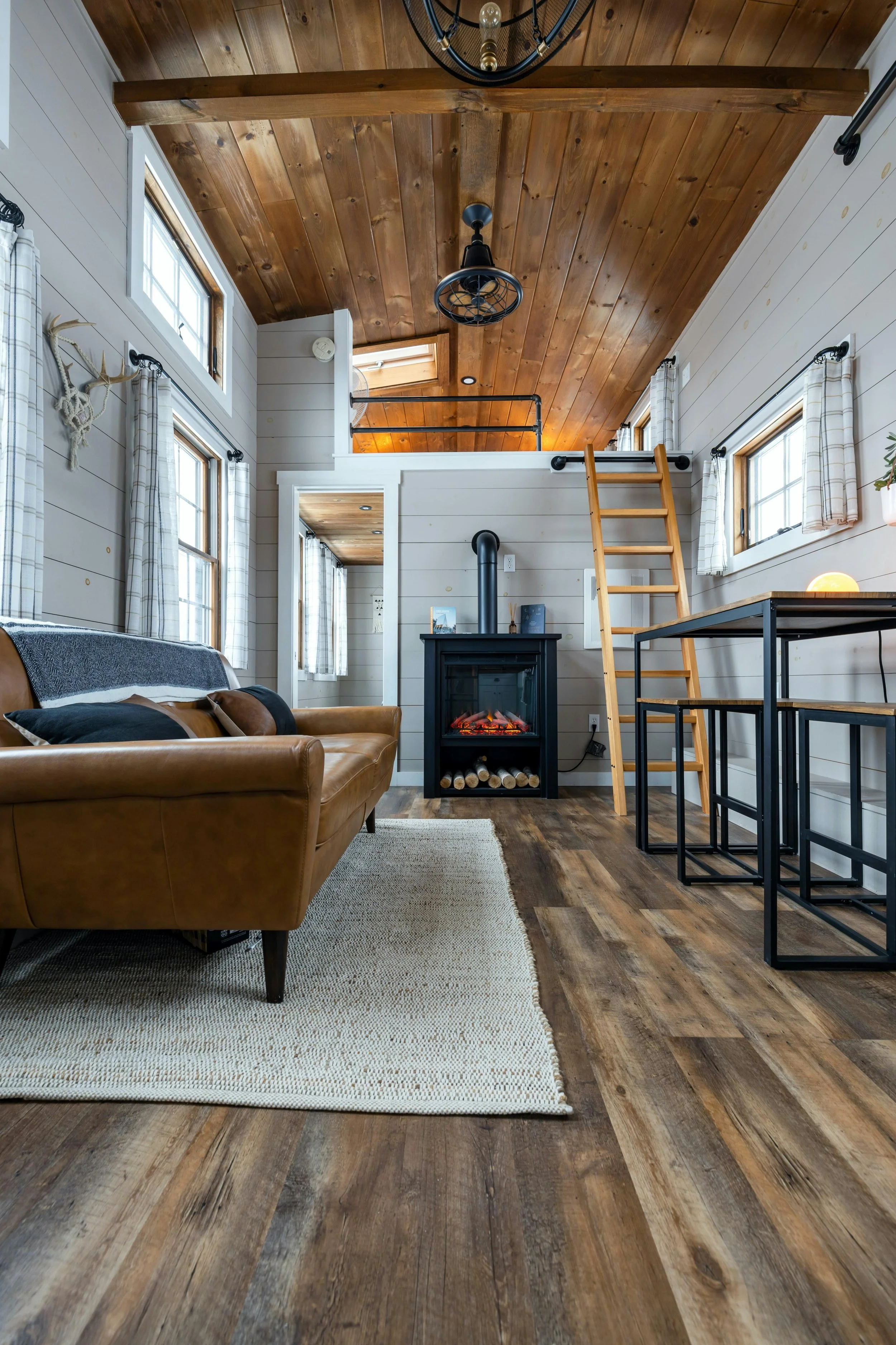 Interior of a cozy ADU living space with a wooden ceiling and floors, white shiplap walls, large windows with plaid curtains, a leather sofa, a small electric fireplace, a ladder leading to a loft.