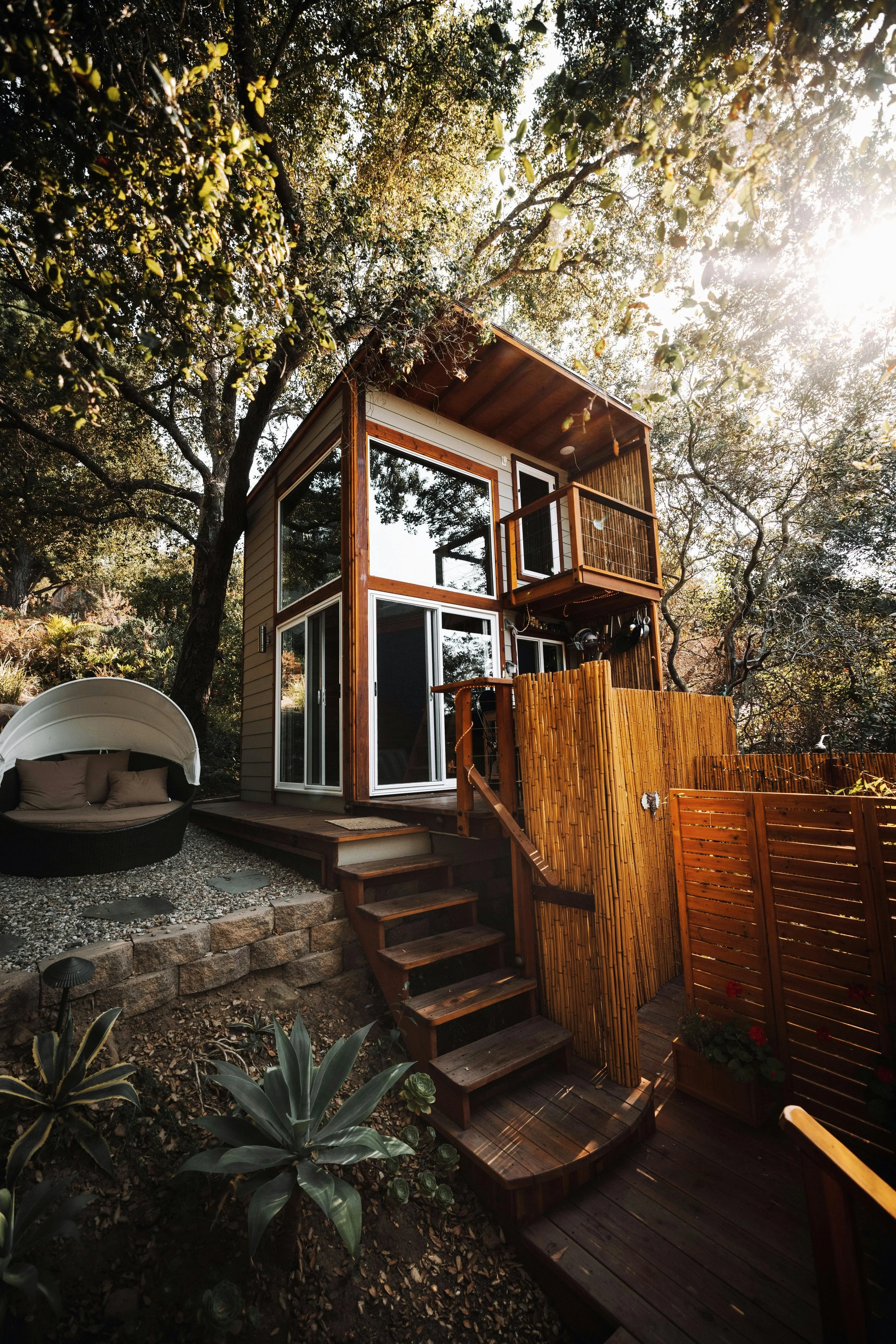 A small two-story ADU with large windows and a wooden balcony nearby surrounded by trees and plants.