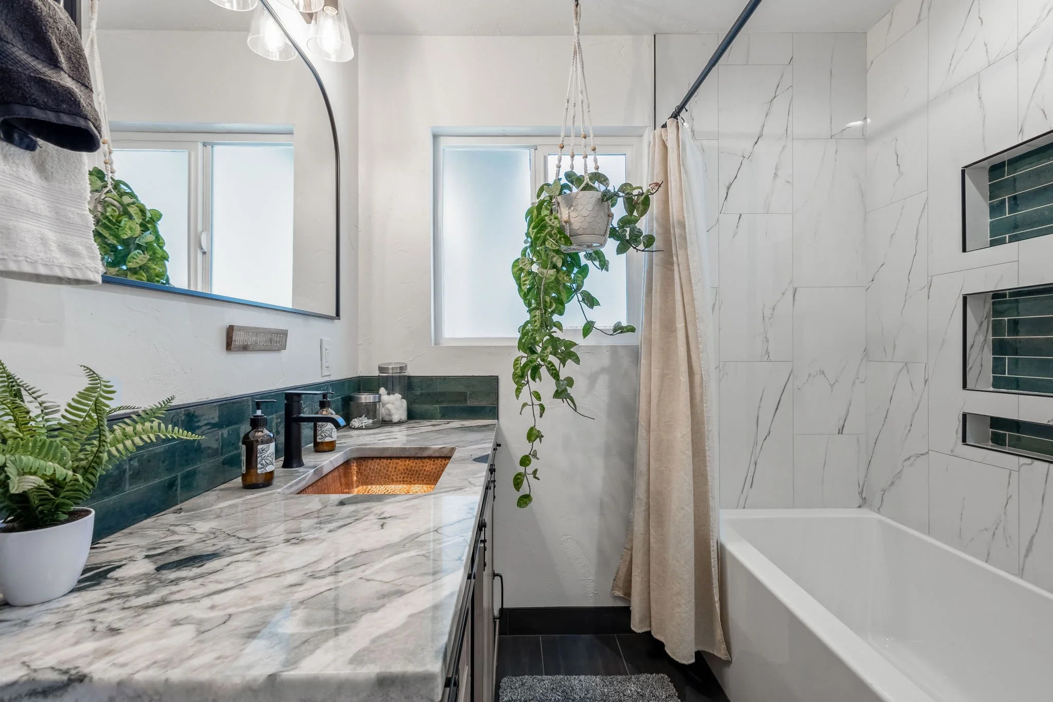 Modern bathroom with marble countertops, green tiled accents, hanging plants, and a bathtub with a shower curtain. There's a large mirror above the sink and windows letting in natural light.