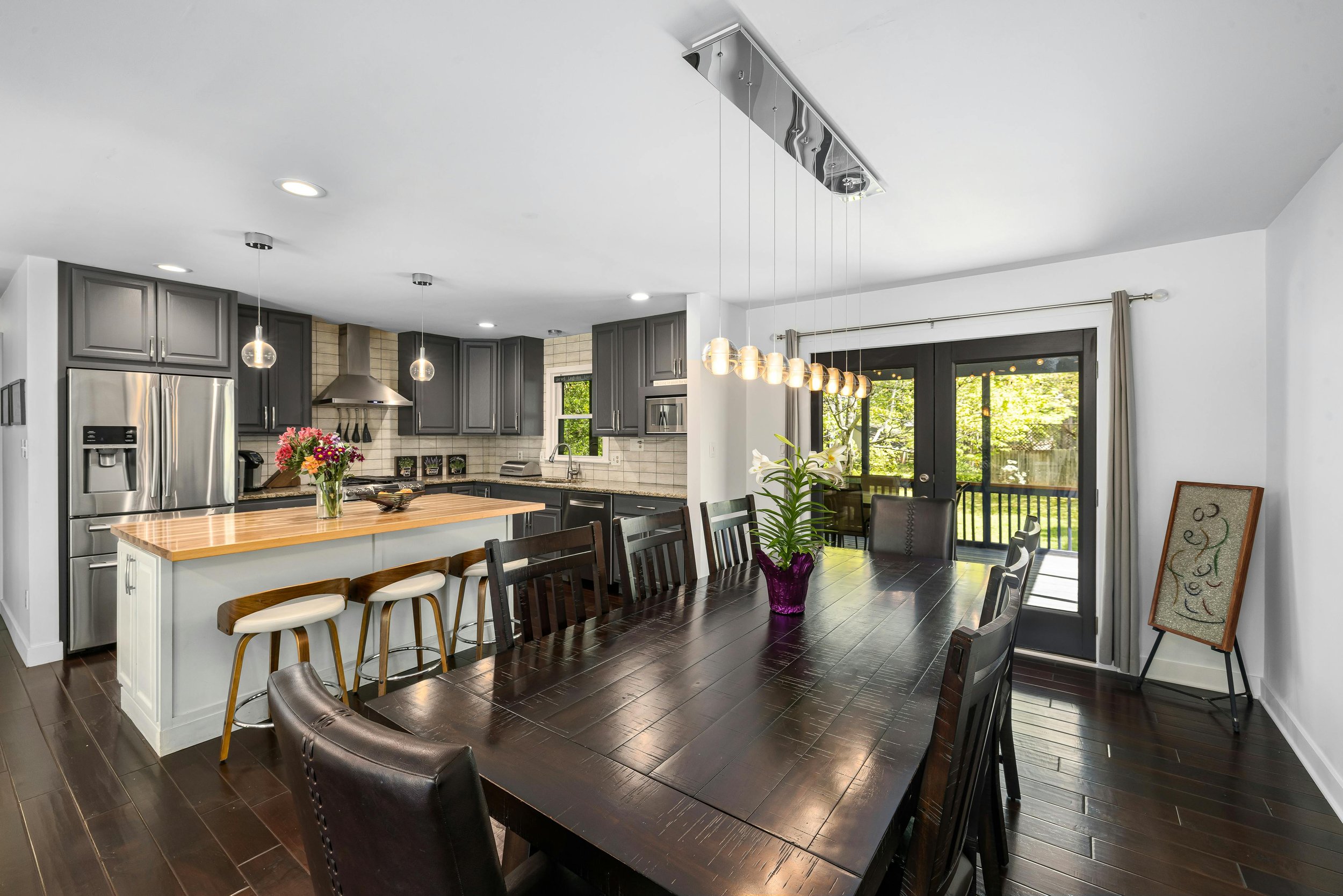 Modern kitchen with gray cabinets, stainless steel appliances, a center island with a wooden top, and a dining table with chairs. There are pendant lights and a sliding glass door leading to an outdoor area with trees visible.
