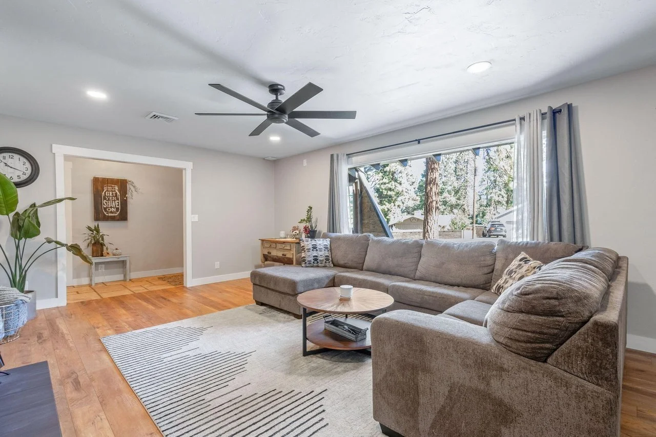 Living room with gray sectional sofa, wooden coffee table, area rug, large window with curtains, ceiling fan, and hardwood floors.
