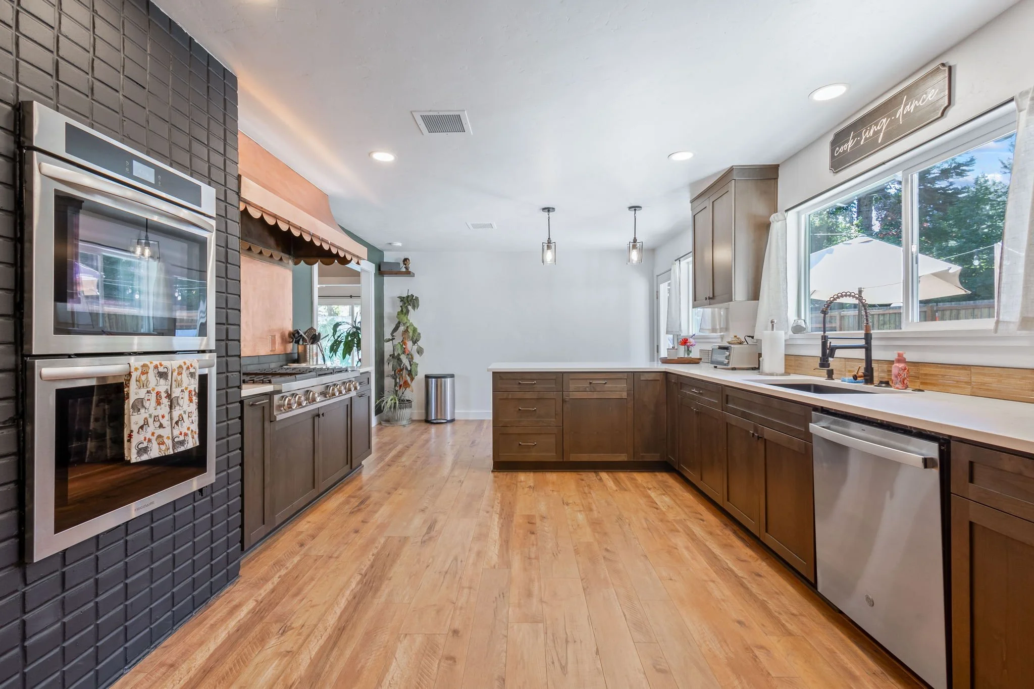 Kitchen with wooden cabinets, stainless steel appliances, a window with white curtains, and light wood floors. Decorative sign reading 'cook. sing. dance' is above the window.