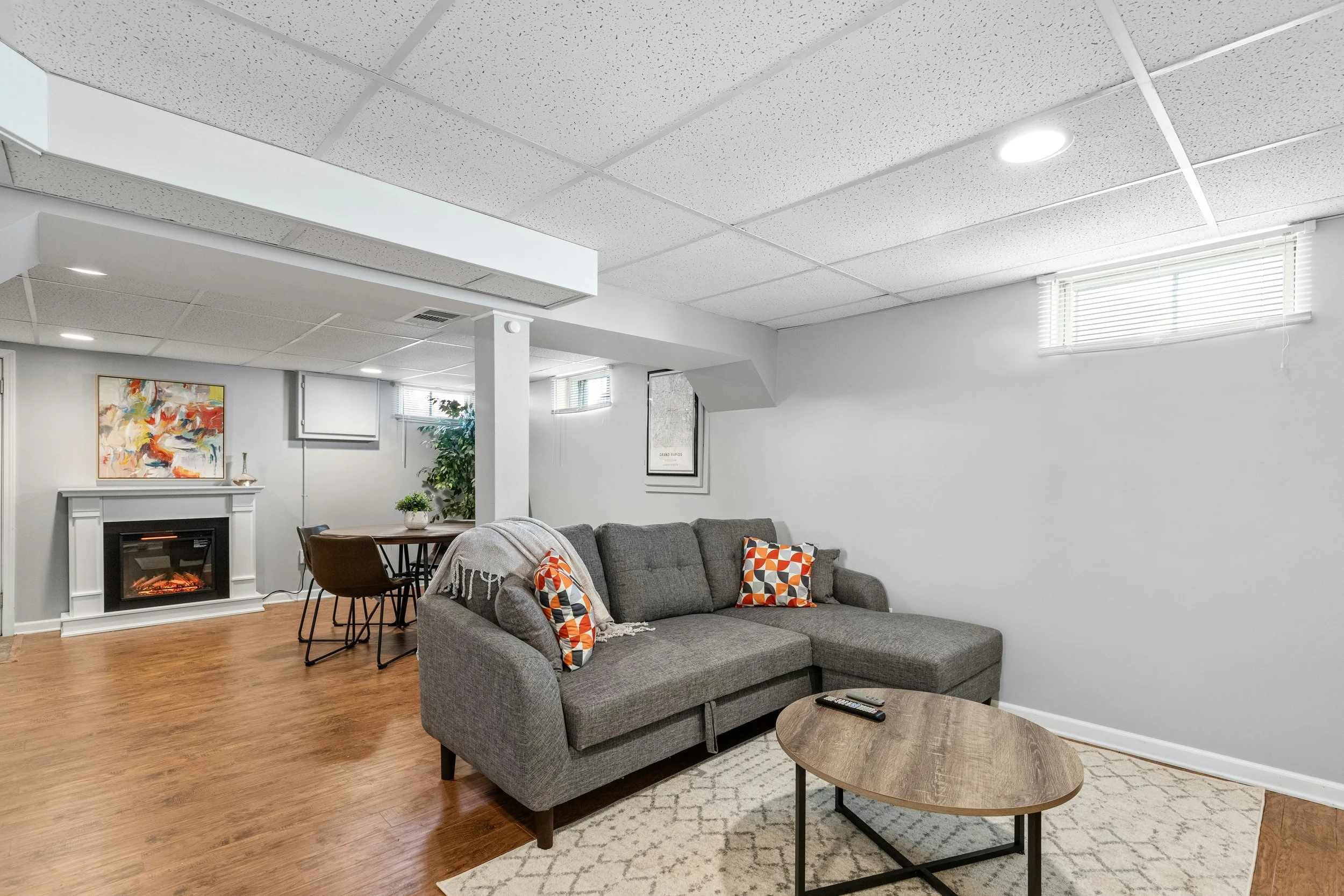 Updated basement with gray sectional sofa, decorative pillows, coffee table, fireplace, artwork, plants, and ceiling lights.