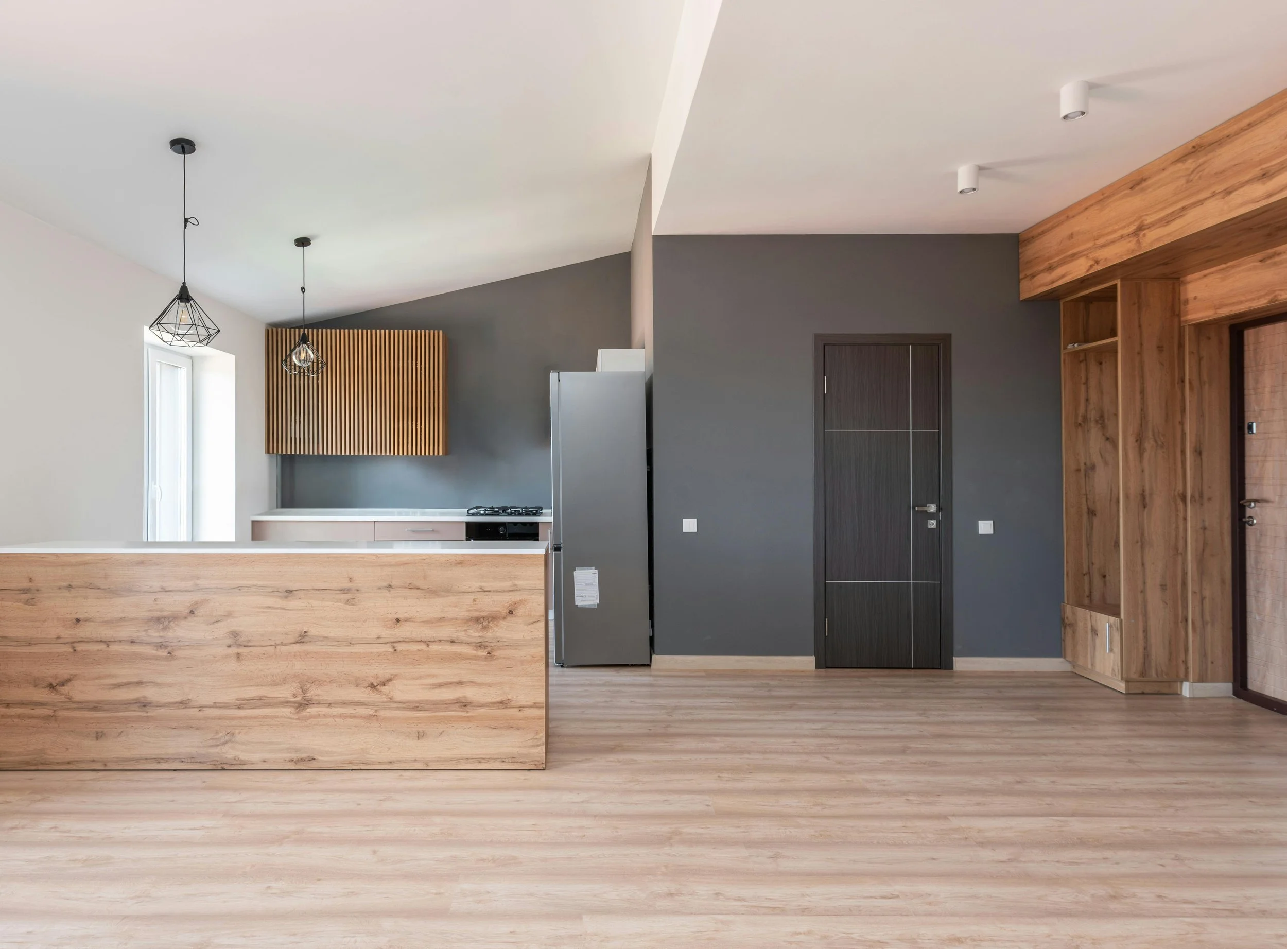A modern apartment with wooden cabinets, stainless steel appliances, including a refrigerator, and a gray color scheme on the walls and door. There are pendant lights hanging from the ceiling and wood flooring.