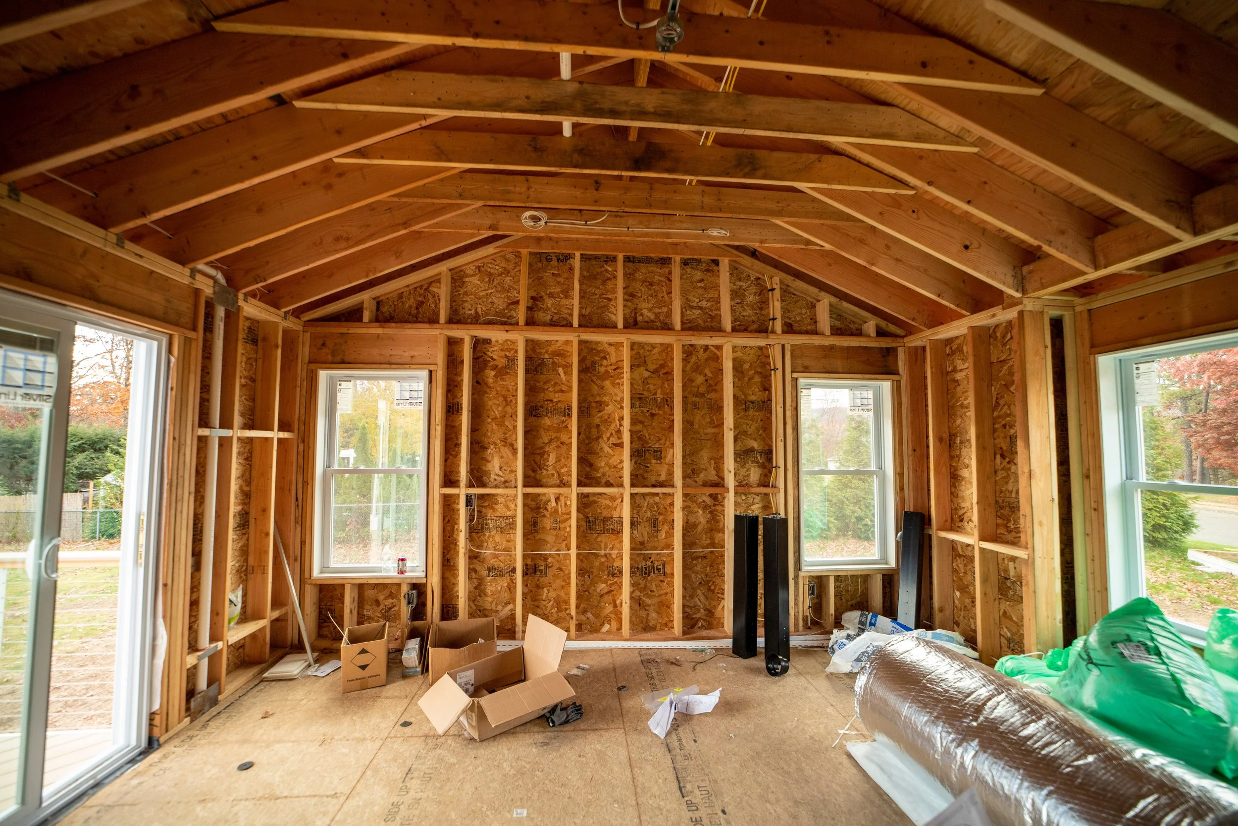 Interior view of a home addition under construction with exposed wooden framing, windows, and construction materials on the floor.