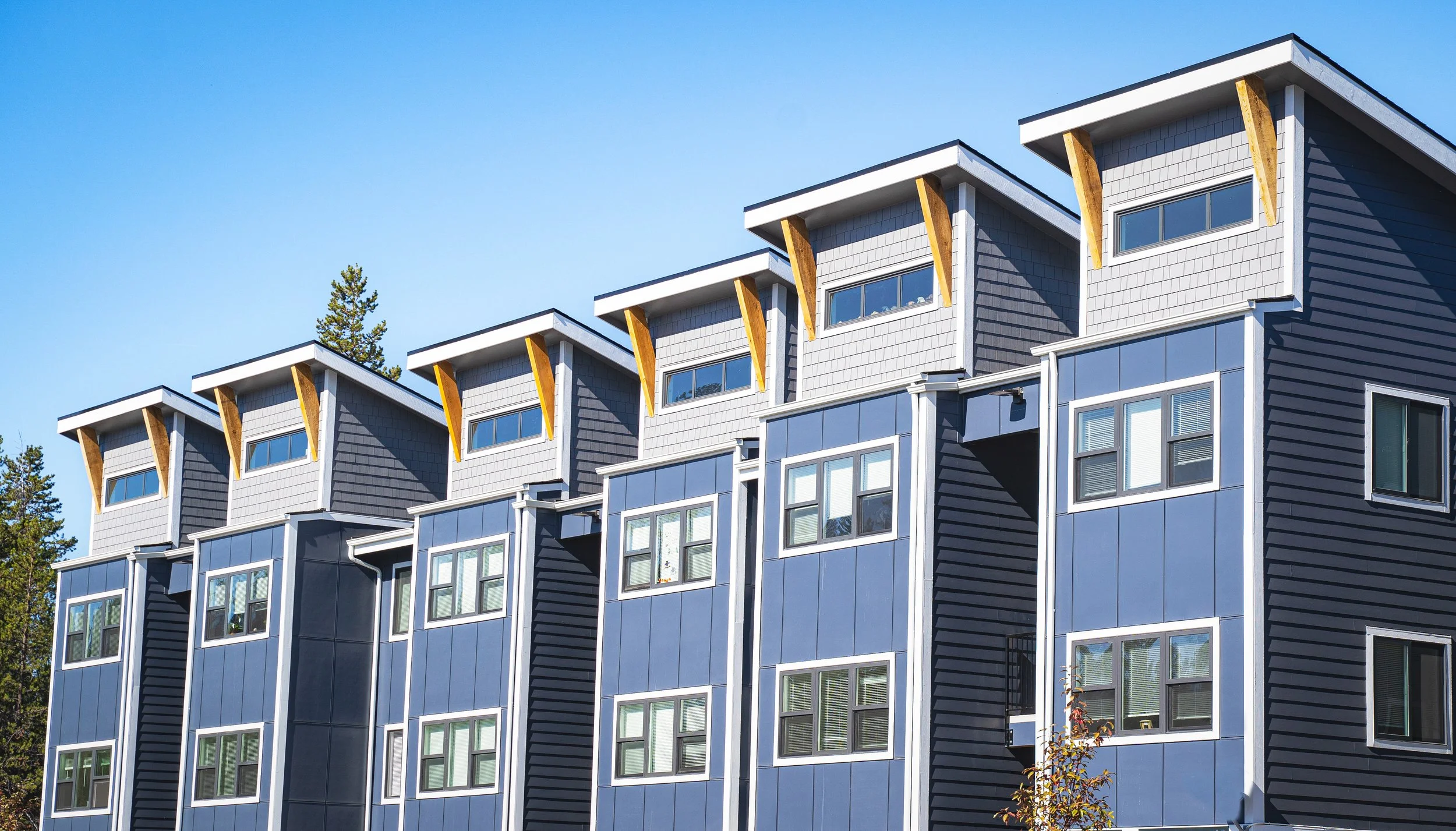 Modern multi-story apartment building with blue siding, large windows, and wooden roof supports, under a clear blue sky.