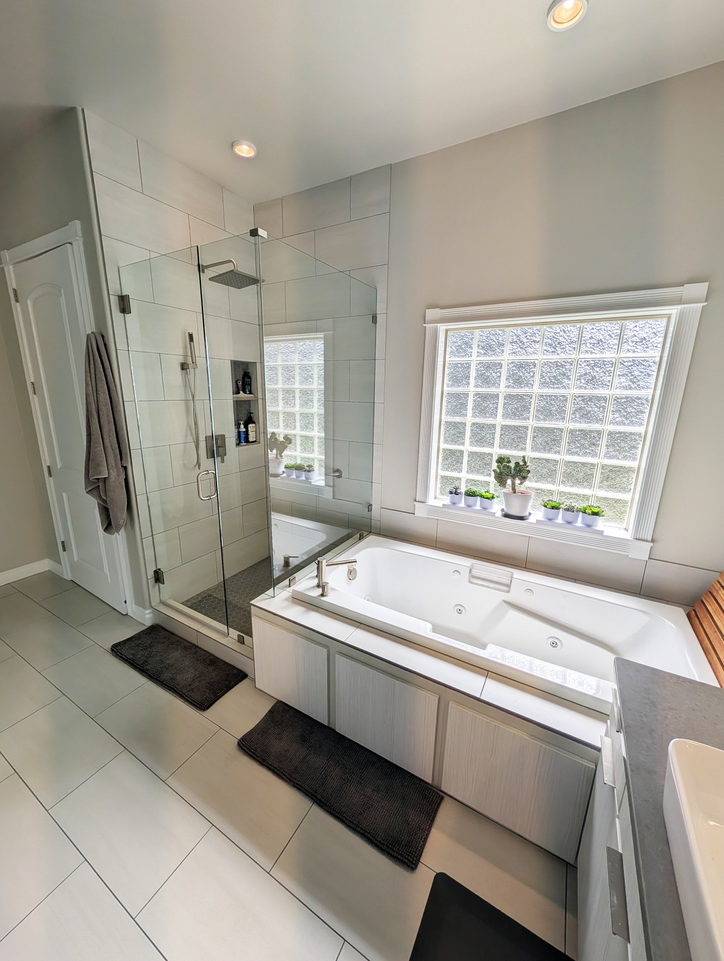 A modern bathroom featuring a glass-enclosed shower with a matte black showerhead and hardware, a built-in shelf with toiletries, a large bathtub with jets, a window with frosted glass blocks topped with white trim, and a surface with potted plants, all with white tiled floor and walls painted in light neutral colors.