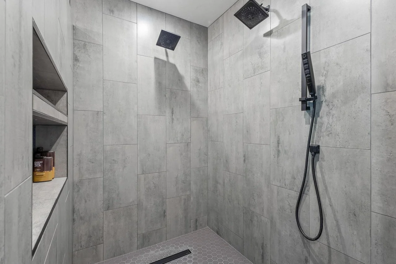 Modern shower with gray tiled walls, two overhead rainfall shower heads, and a handheld shower wand on a sliding bar, with built-in corner shelves.