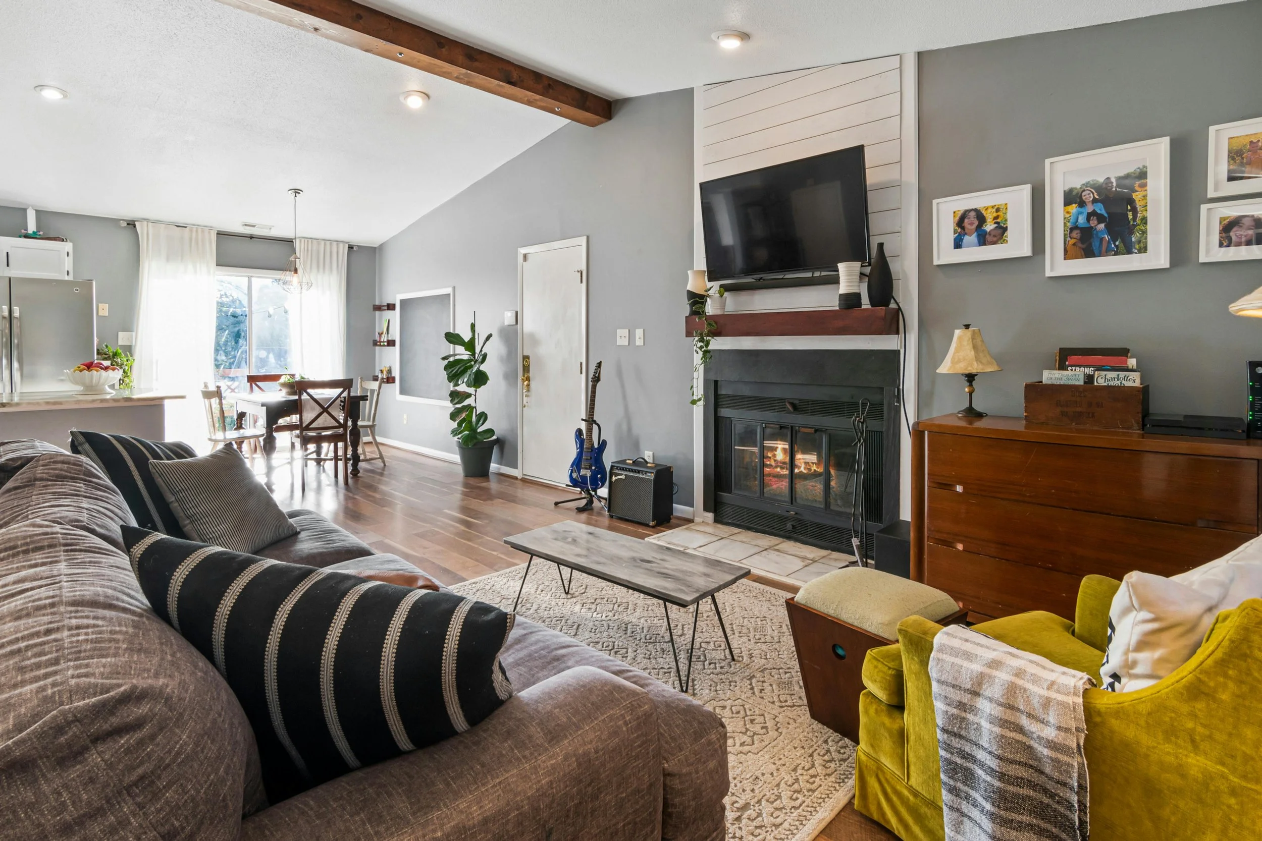 Living room with gray walls, a stone fireplace with a mounted TV, framed family photos, a green velvet armchair with a blanket, a gray sofa with striped pillows, a wooden side table, a patterned rug, a potted plant, and a dining area in the background with natural light from a large window.