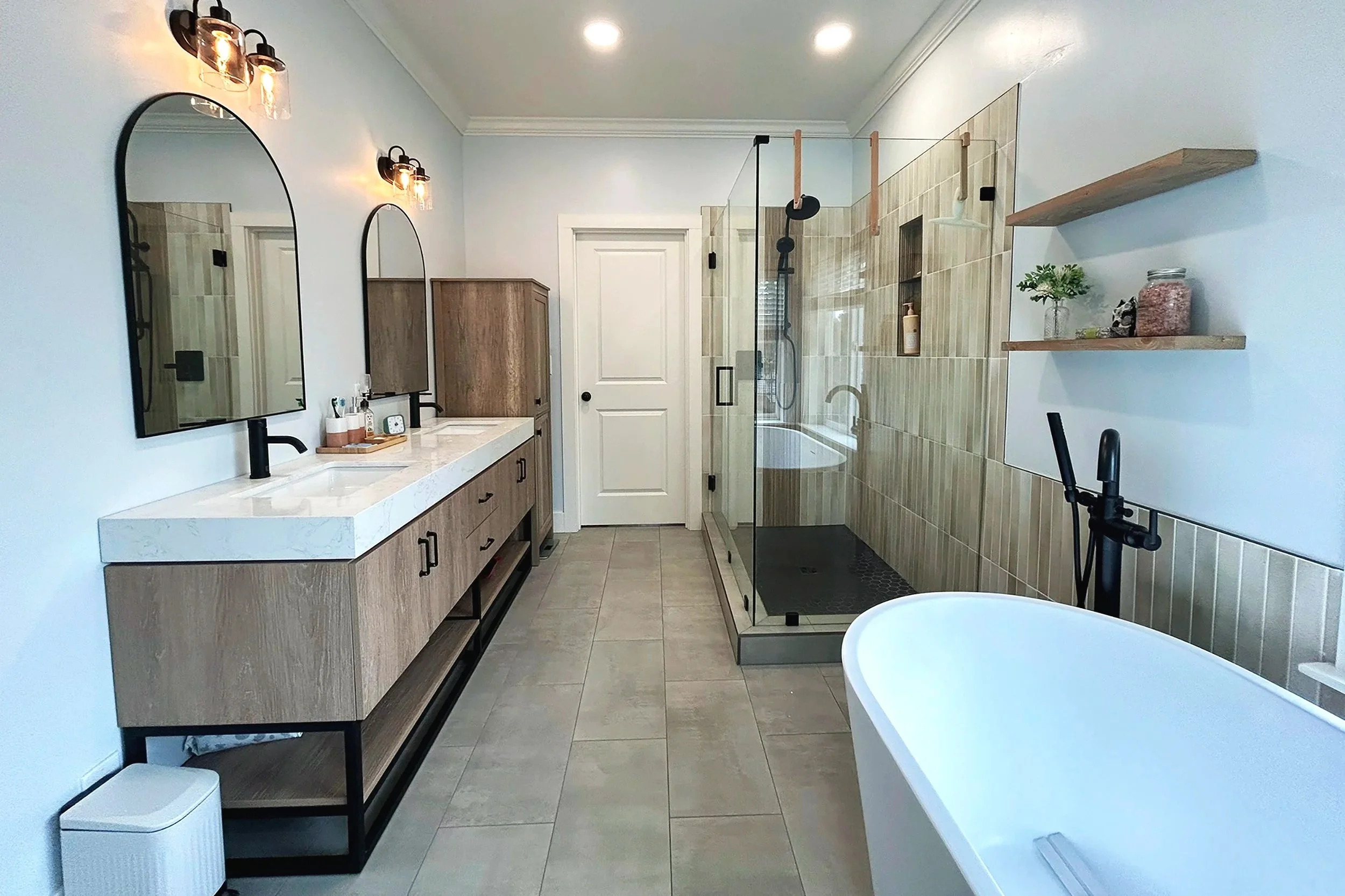 Modern bathroom with double vanity, round mirrors, tan cabinetry, two black wall-mounted light fixtures, glass-enclosed shower with tiled walls, white bathtub, floating wooden shelves with decorative items, and beige tiled flooring.