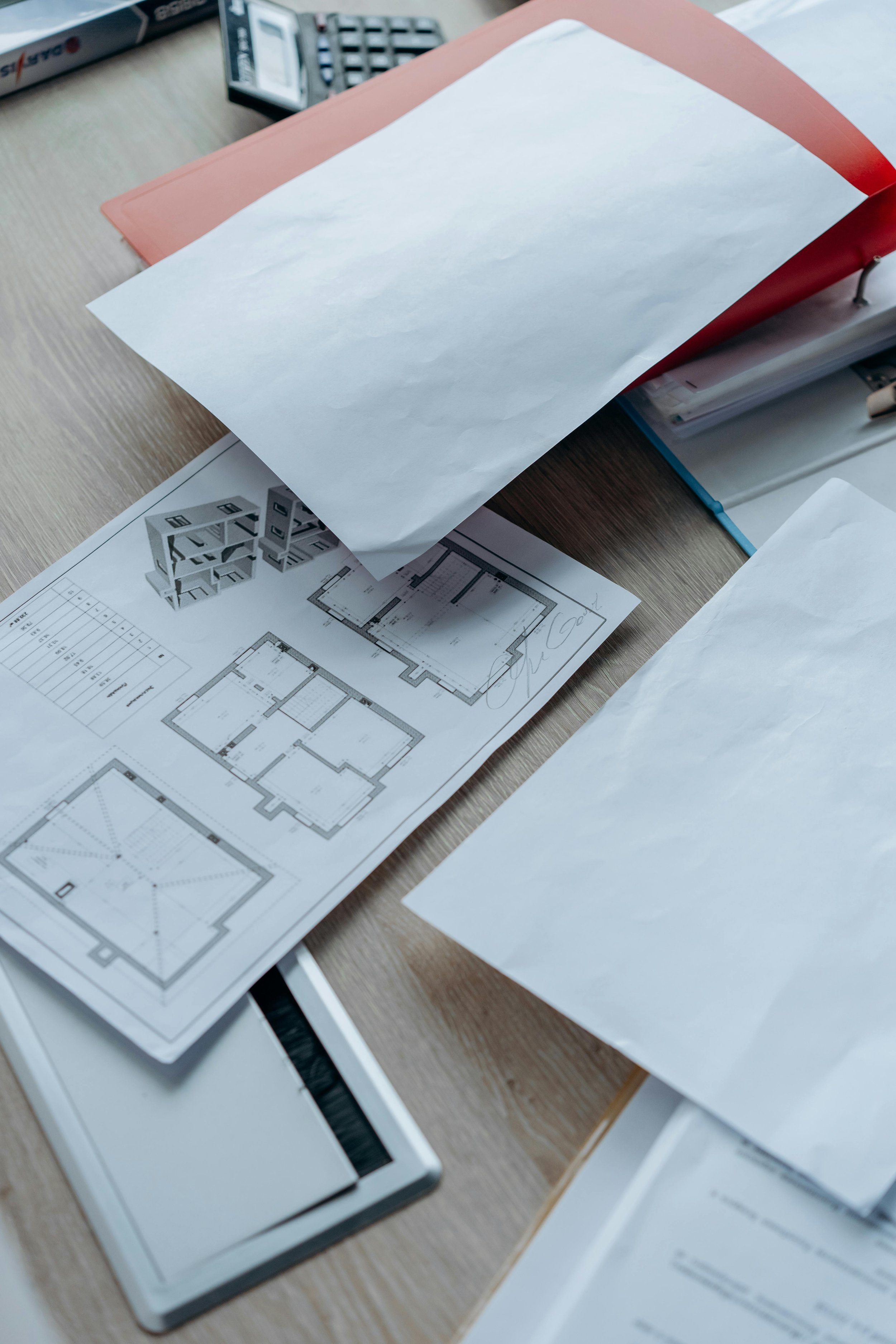 Architectural blueprints, design plans, and drafting tools on a wooden desk, including a scale ruler, a red binder, and handwritten notes.