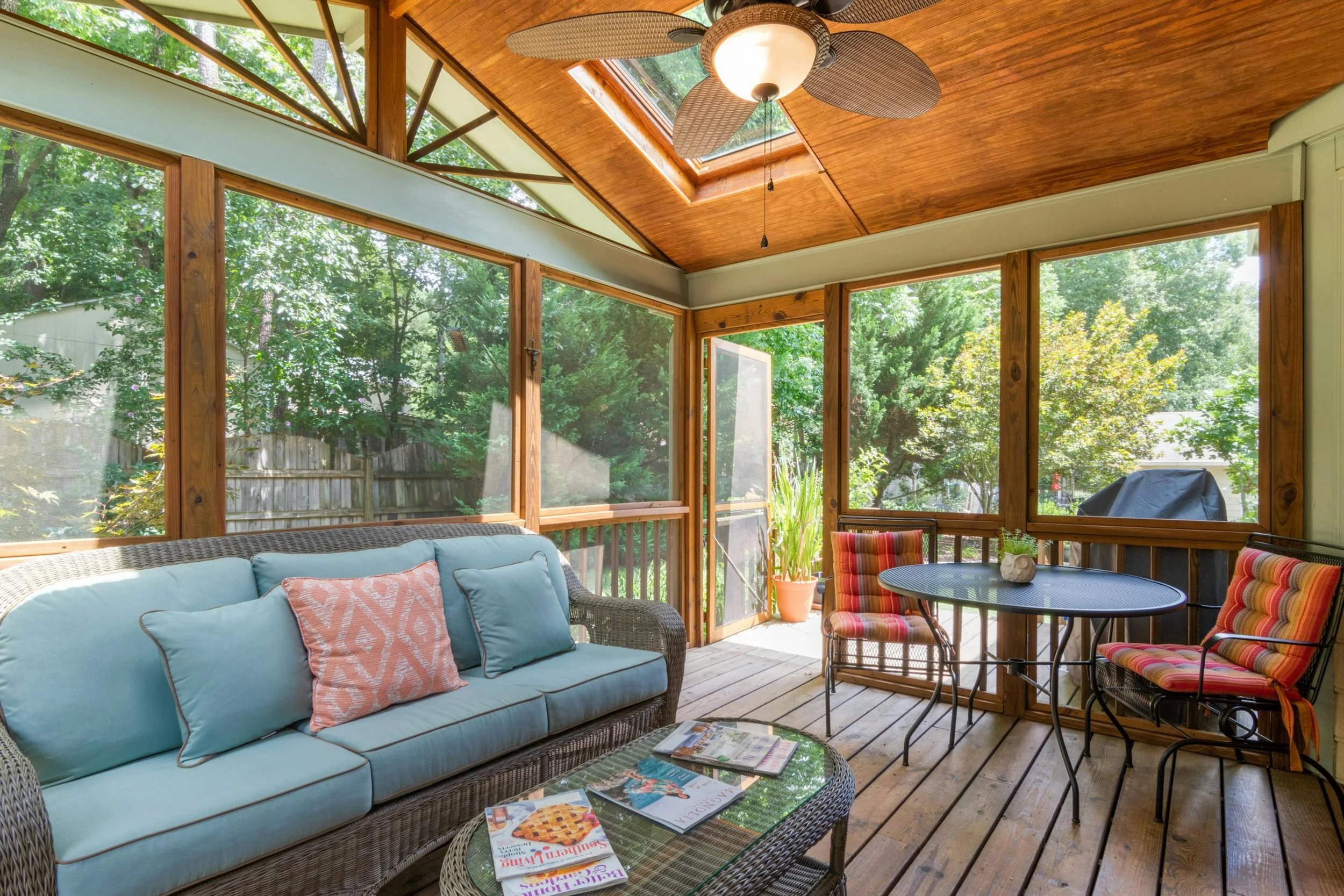 A screened porch addition with wooden ceiling and support beams, large windows showing green trees outside, a ceiling fan, a light fixture, a sofa with cushions, a round table with magazines, and additional chairs with colorful cushions.