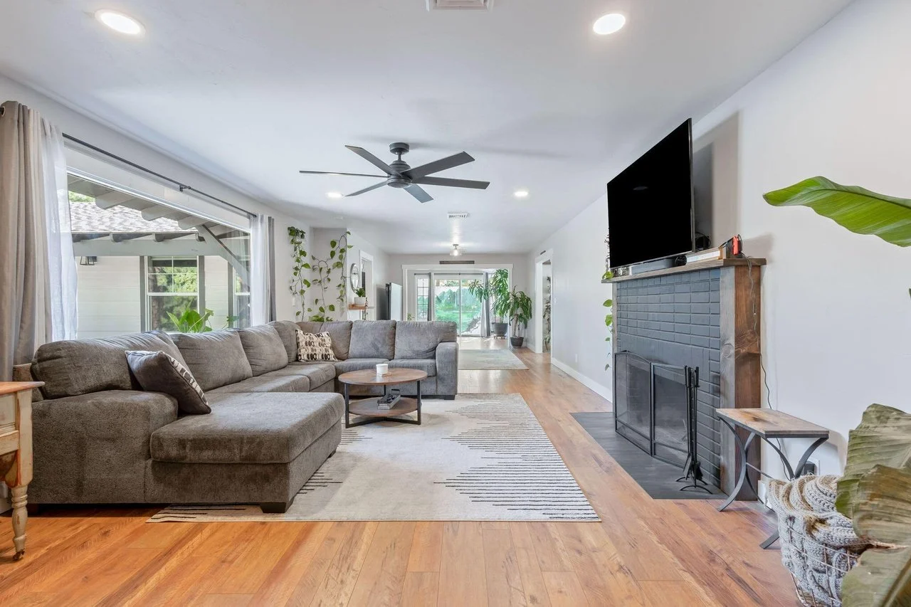 Living room with gray sectional sofa, wooden coffee table, and fireplace with a mounted TV, hardwood floors, large window with curtains, and potted plants, leading to a sunny area with a glass door.
