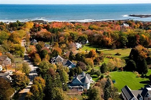 Aerial view of a neighborhood with colorful autumn trees, houses, and a golf course near the shoreline of an ocean or large body of water.