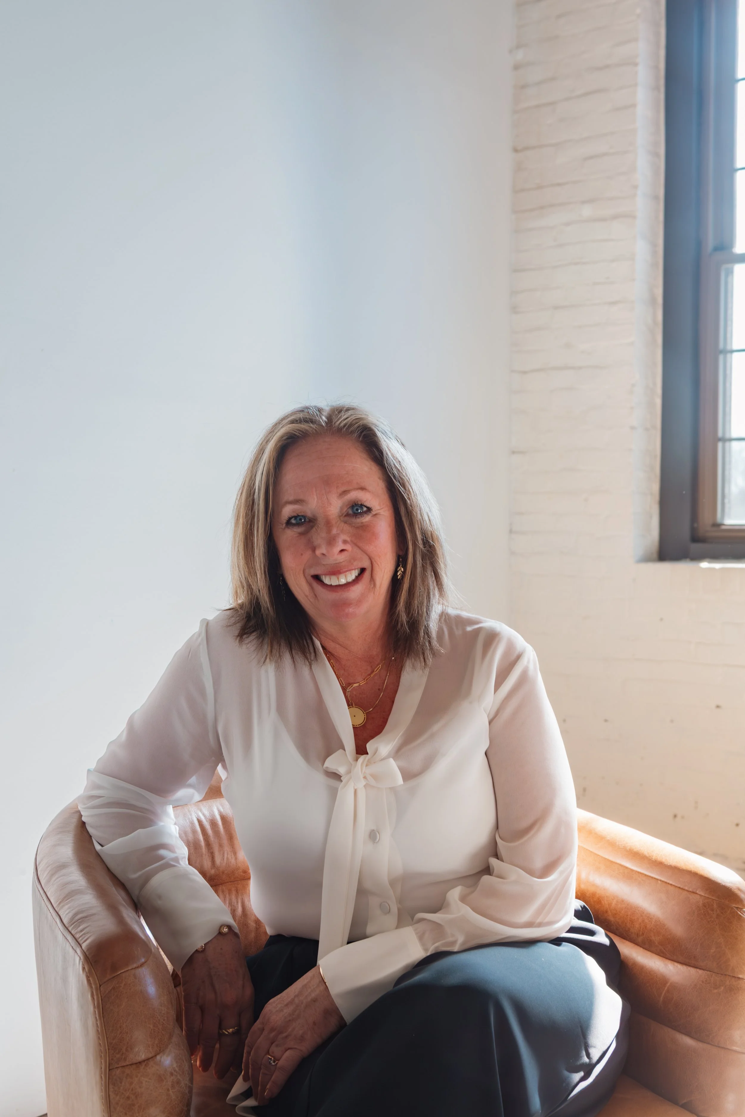 A woman sitting on a tan leather armchair in a room with white walls and a large window. She has shoulder-length hair, wearing a white blouse with a bow, gold jewelry, and dark pants. She is smiling and looking at the camera.