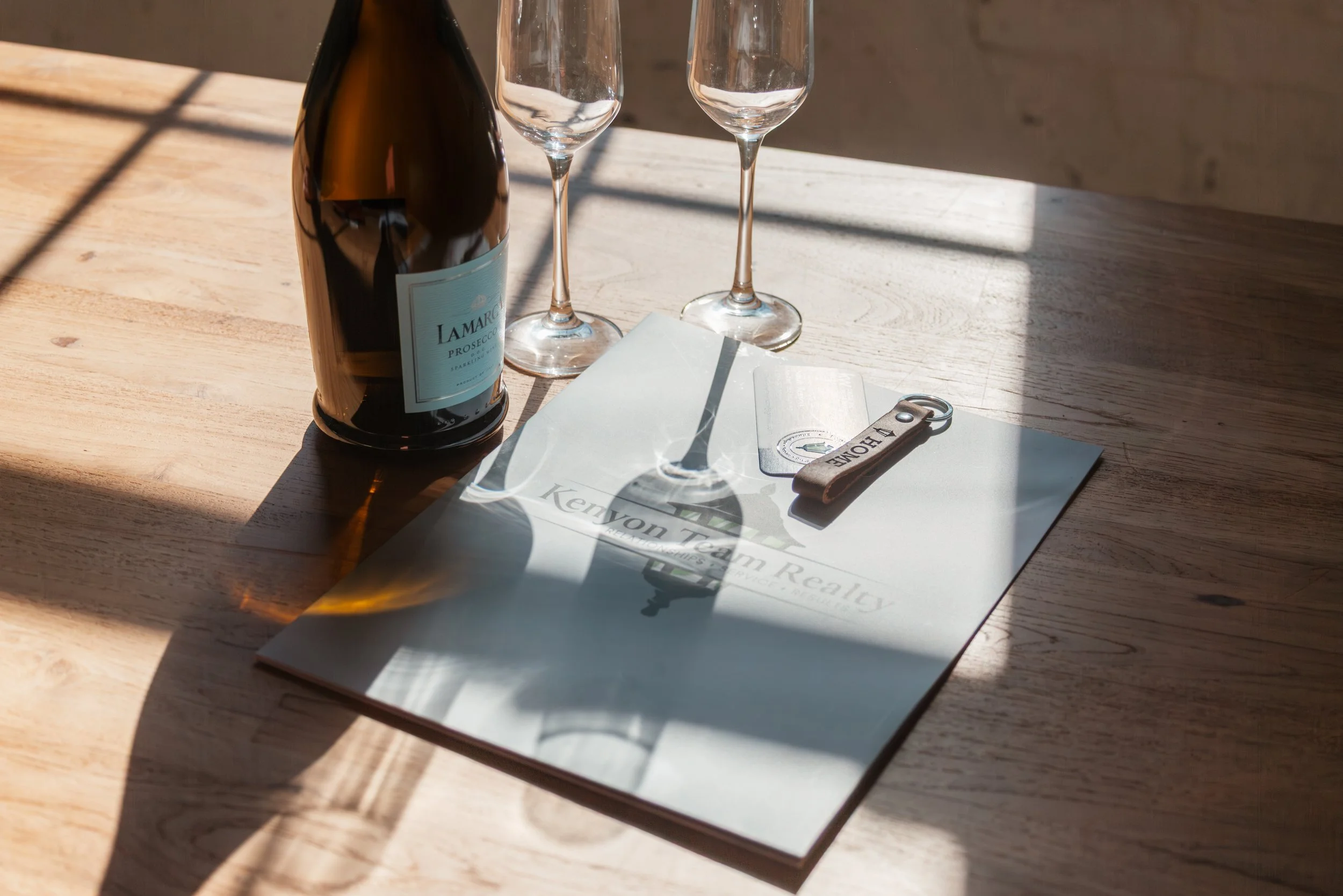 A bottle of Lamaro Prosecco, two empty champagne glasses, a keychain with the word 'HOME' on it, and a folded paper on a wooden table, with sunlight casting shadows.