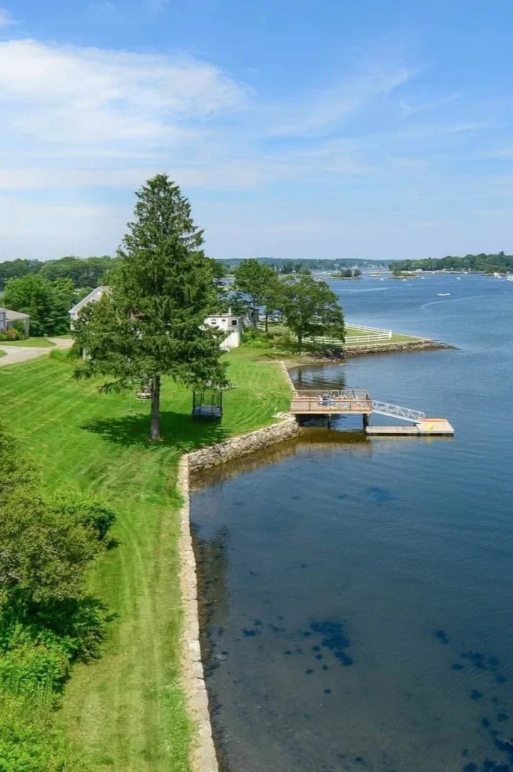 View of a lakeside property with a grassy yard, several trees, a wooden dock with a ramp, and a view of the water with boats in the distance.