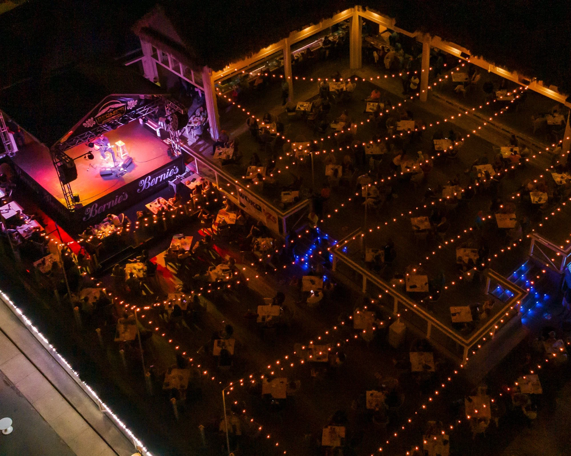 Nighttime scene of an open-air restaurant with string lights, tables, and people dining. A stage with musical instruments and performers is visible in the lower part of the image.