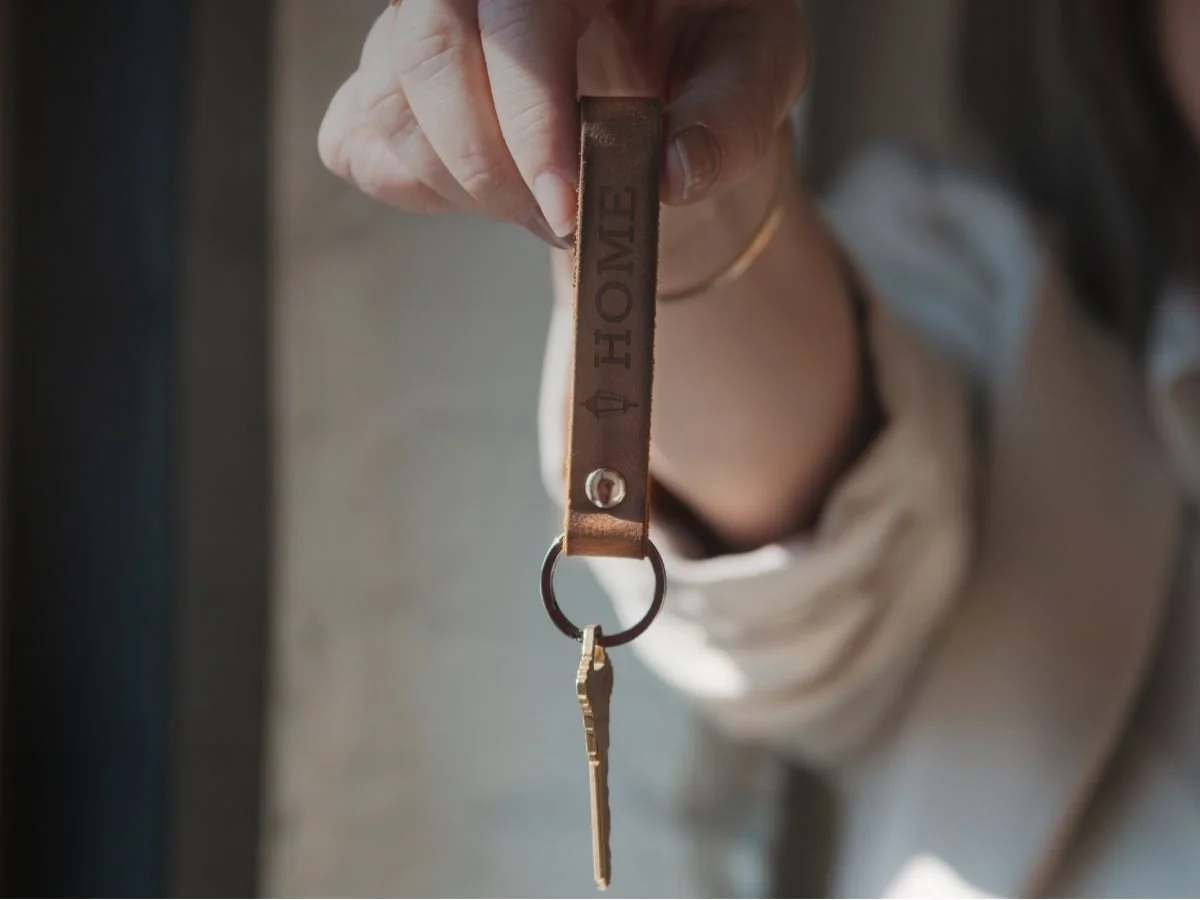 Close-up of a person's hand holding a leather keychain with the word 'HOME' engraved on it, with a key attached.