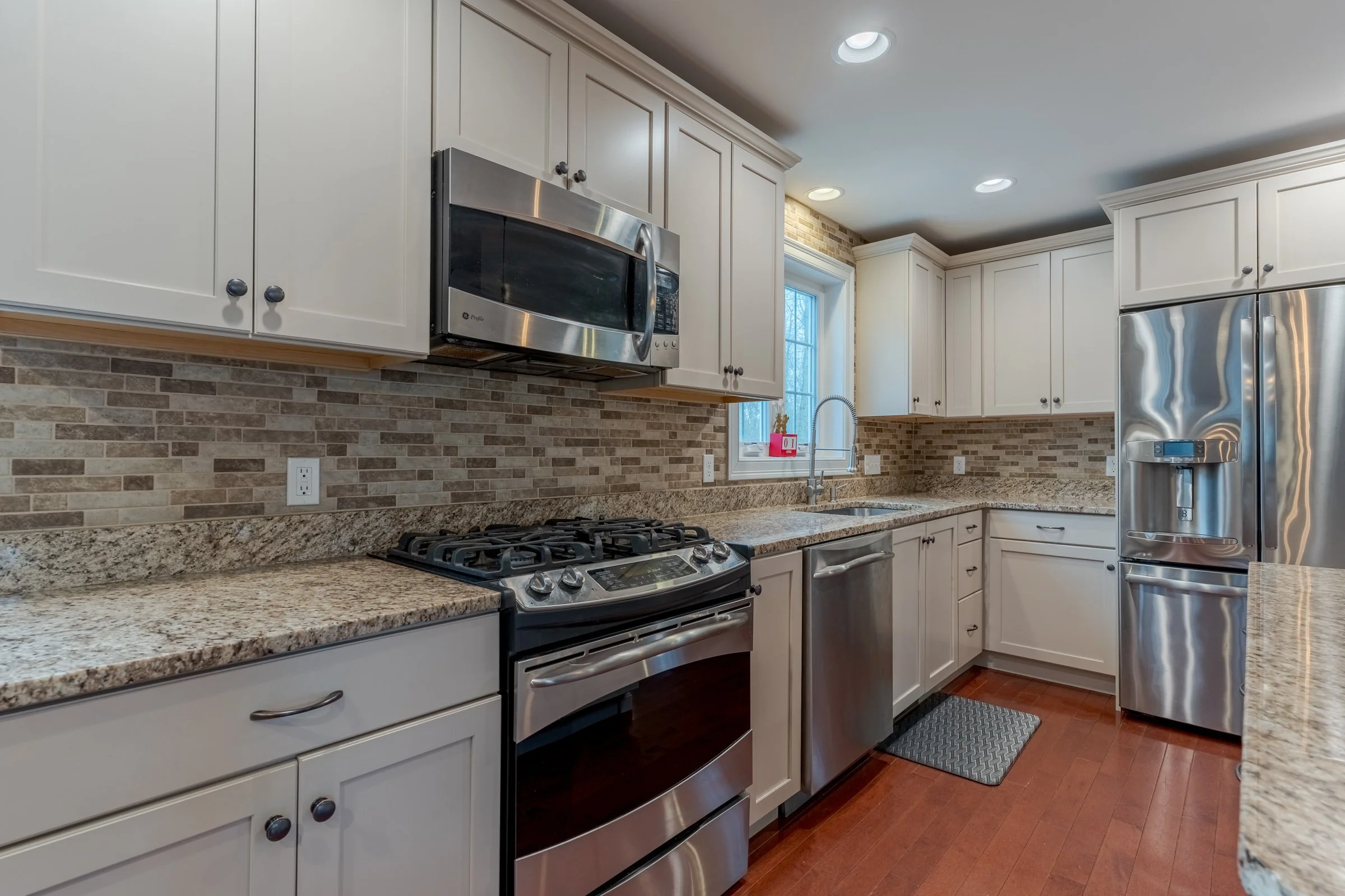 A modern kitchen with white cabinets, a granite countertop, stainless steel appliances including a microwave, oven, and refrigerator. There is a window above the sink, brick backsplash, and recessed lighting on the ceiling.