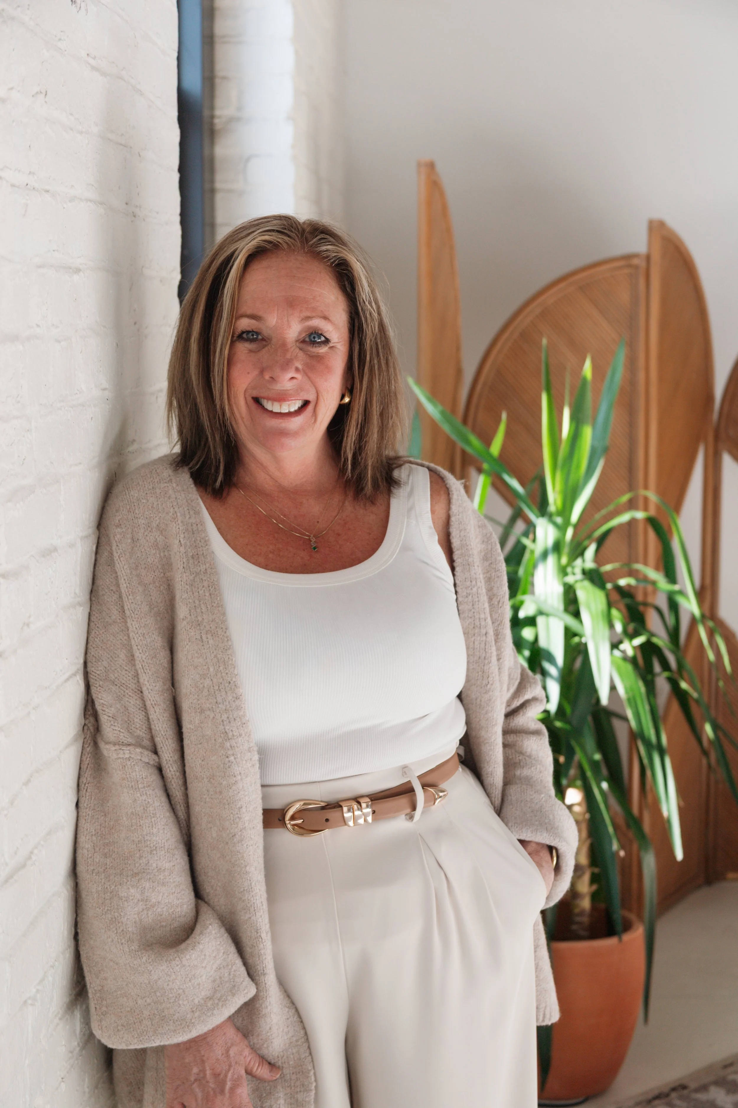 A smiling woman with shoulder-length light brown hair and blue eyes, wearing a white top, beige cardigan, and cream-colored pants with a beige belt, standing indoors next to a white brick wall and green potted plant.