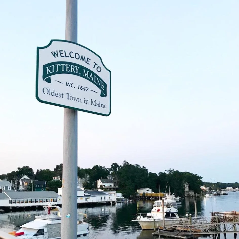Welcome sign for Kittery, Maine, showcasing the town's pier with boats docked along the water and houses in the background under clear sky.