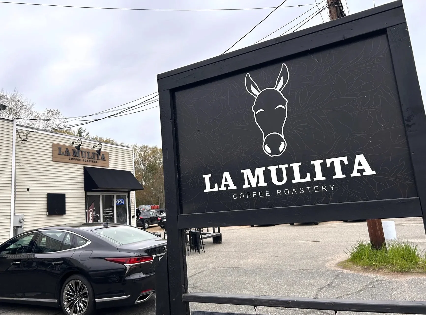 Sign for La Mulita Coffee Roastery with a cartoon donkey's head above the text. The sign is outside near a parking lot, with a white building and cars in the background.