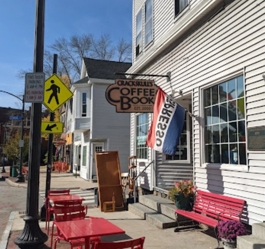Street view of Crackskully's Coffee Book storefront with outdoor seating, a red bench, potted flowers, and a sign hanging above the entrance. A French flag is also visible.