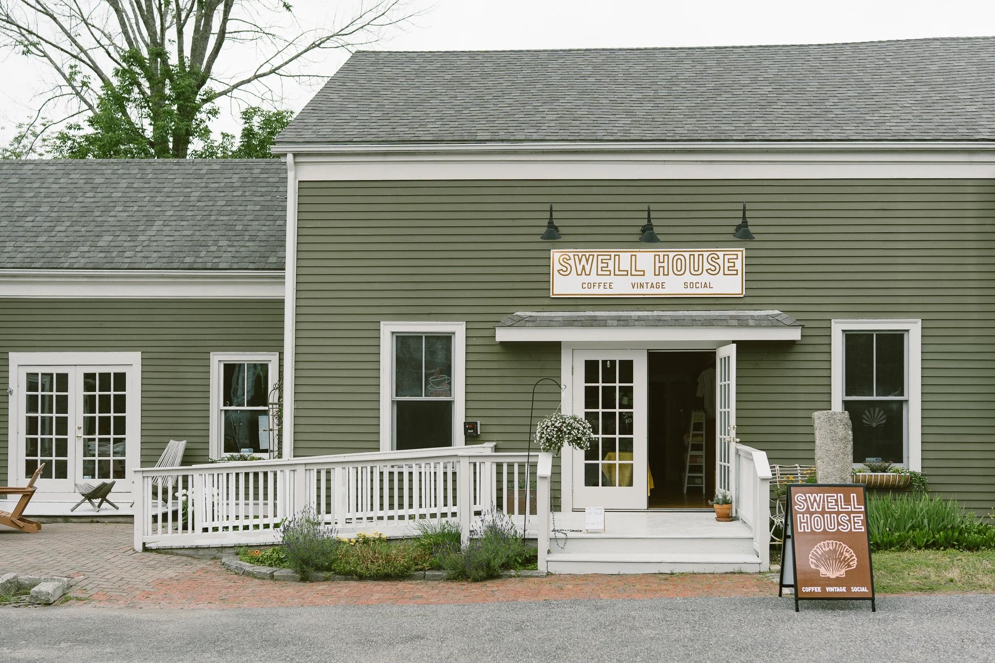 Exterior of a green building with a sign that reads "Swell House" and subtext "Coffee Vintage Social". The building has white-framed windows and a white patio fence in front, with a small flowerpot, lavender plants, and chairs outside.