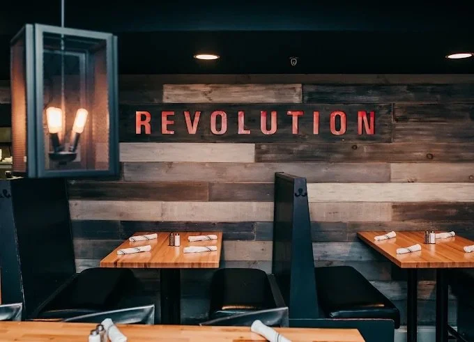 Interior of a restaurant with wooden tables, black chairs, and a wall with the word 'REVOLUTION' displayed in red letters.