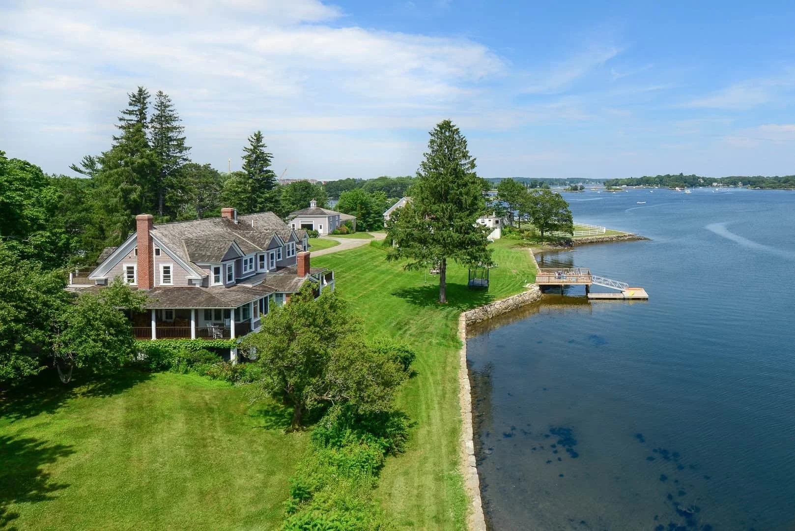Aerial view of a waterfront house with a large porch, surrounded by lush green lawns and trees, overlooking a calm lake with boats and a dock.
