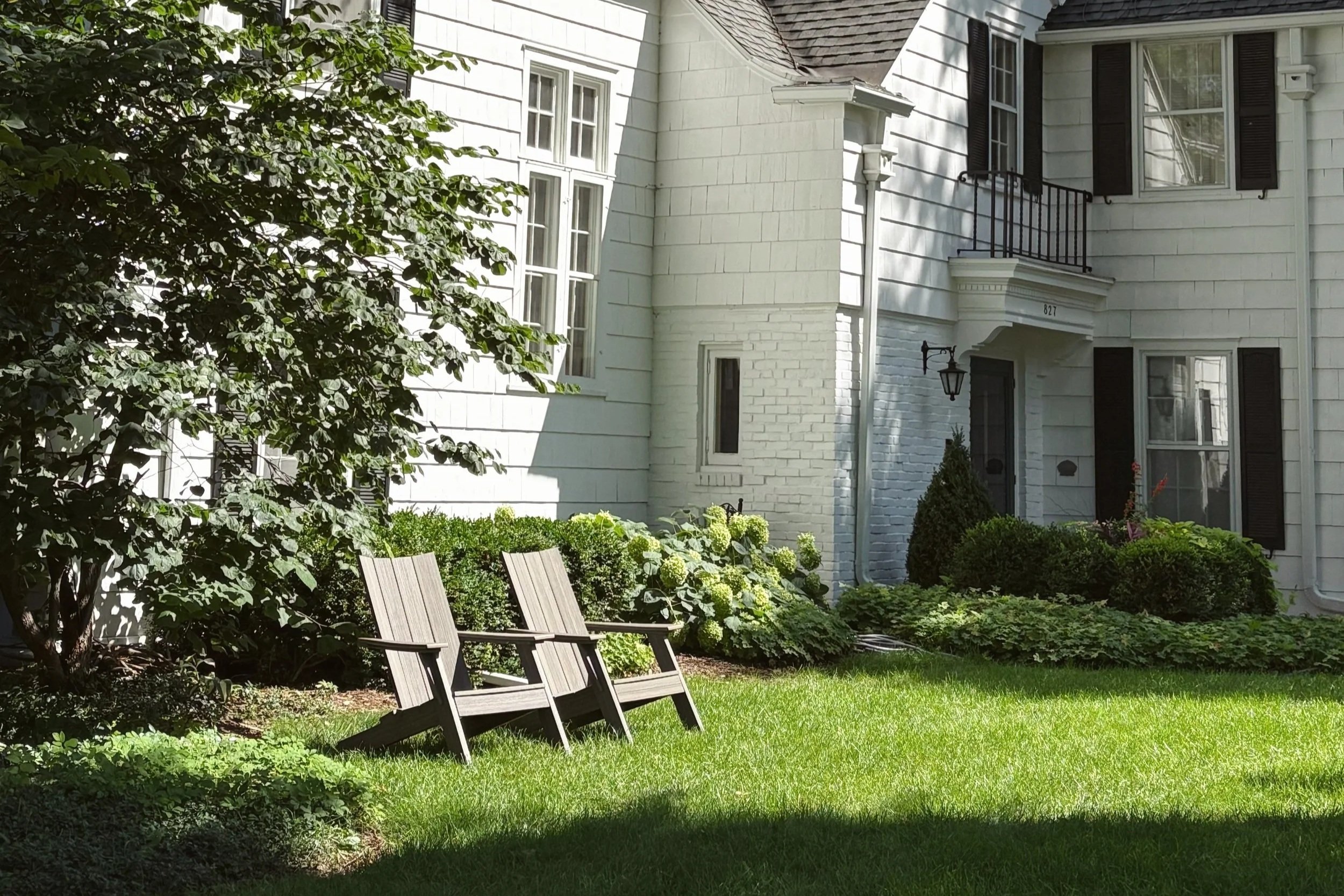 A white house with black shutters, a balcony, and exterior lights. The house is surrounded by a garden with chairs, green grass, bushes, and trees.