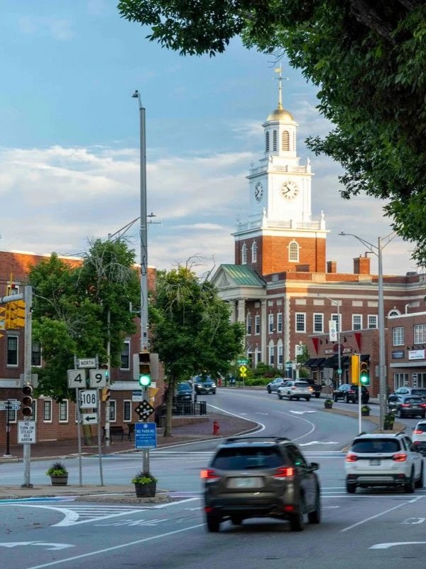 A street corner with cars, traffic lights, and street signs in front of a brick town hall with a clock tower and a gold weather vane, surrounded by trees and buildings under a partly cloudy sky.