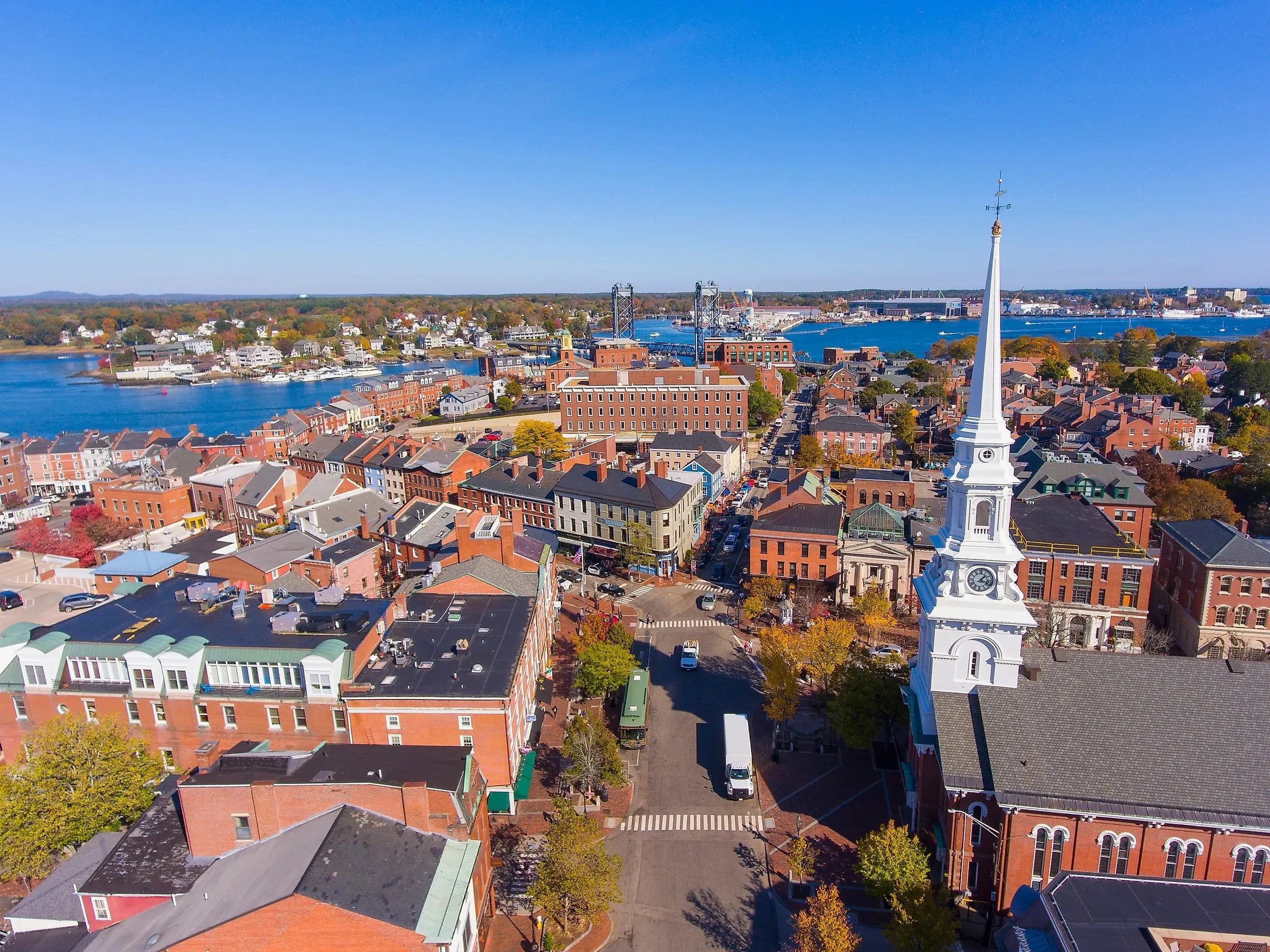 Aerial view of a coastal town with red-brick buildings, waterfront docks, and a large white church steeple with a clock, under a clear blue sky.