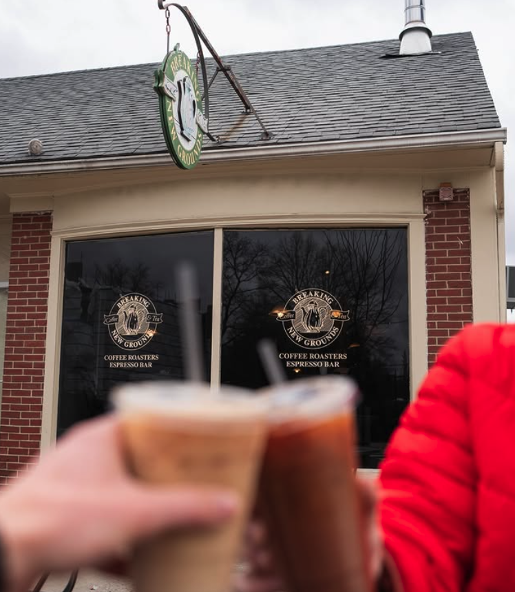 Two people holding cups of coffee outside a coffee shop with a sign that reads "Breaking New Grounds Coffee Roasters Espresso Bar" on the window.