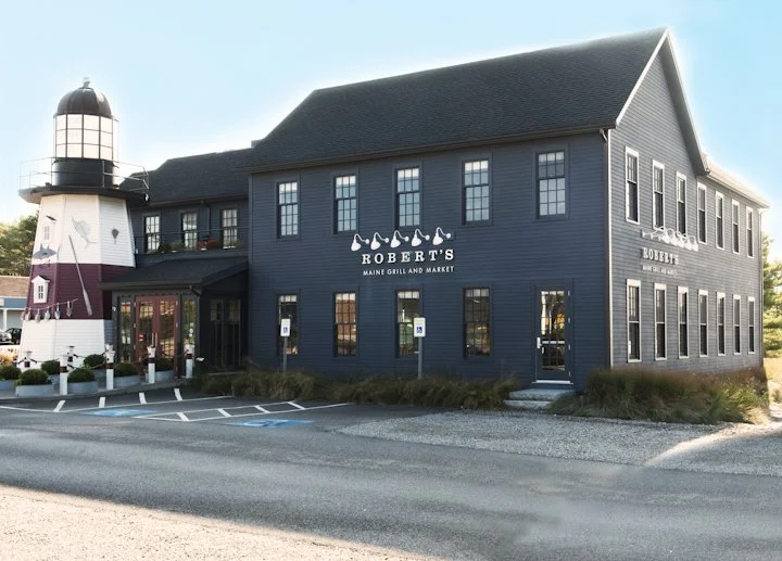 Exterior of Robert's Maine Grill and Market with a lighthouse, dark blue building, and parking lot.