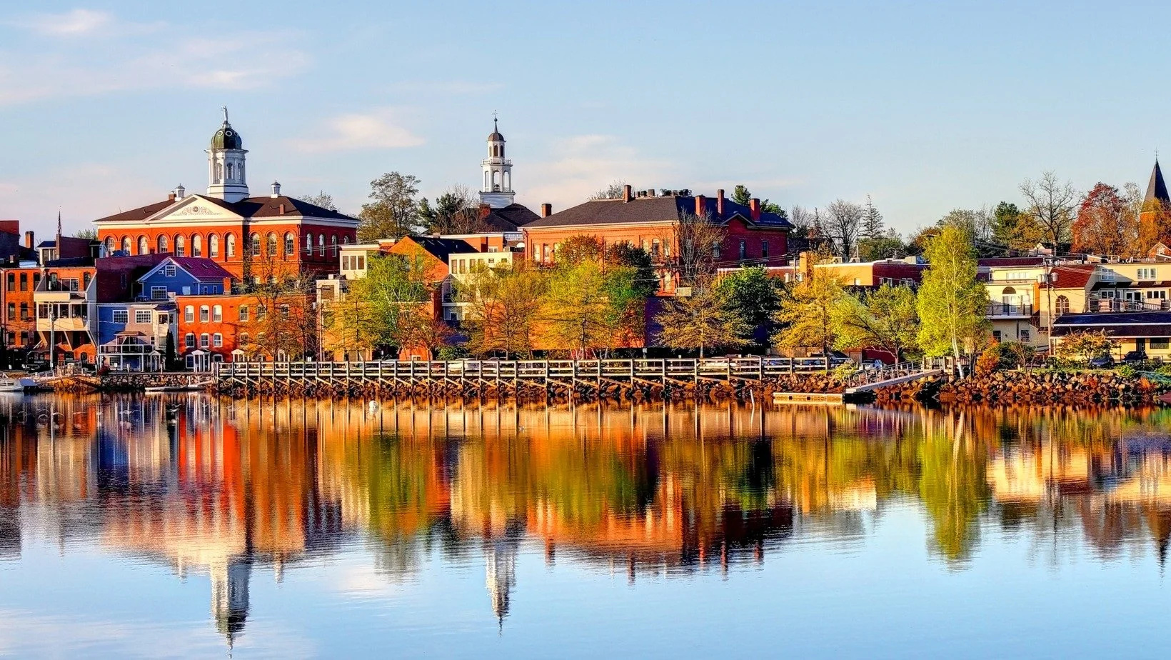 A scenic view of a small town waterfront with colorful buildings, trees, and church steeples reflected in calm water.