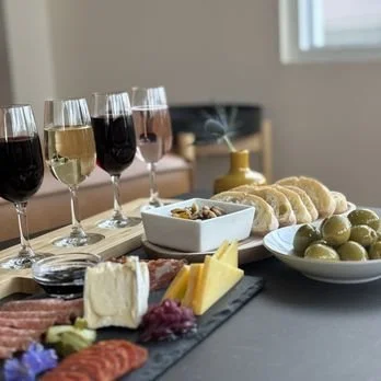 A table with a cheese platter, grapes, sliced bread, and glasses of red, white, and rosé wine in a cozy dining room.
