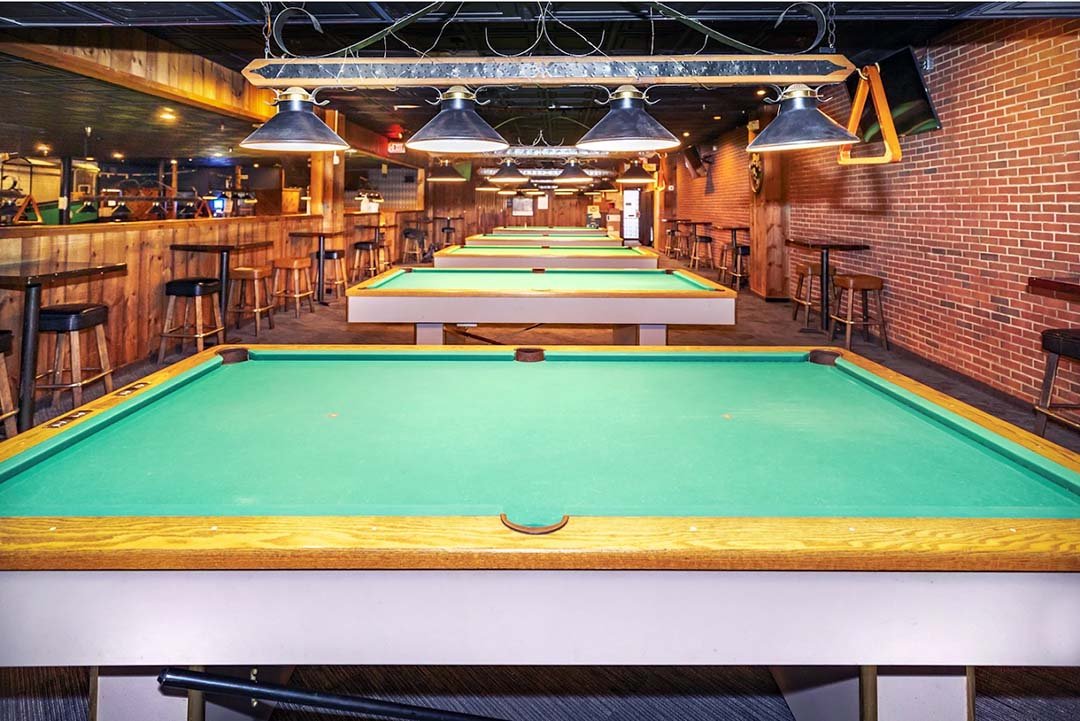 Multiple pool tables lined up in a bar or game room with wooden decor, hanging lamps, and bar stools along the sides.