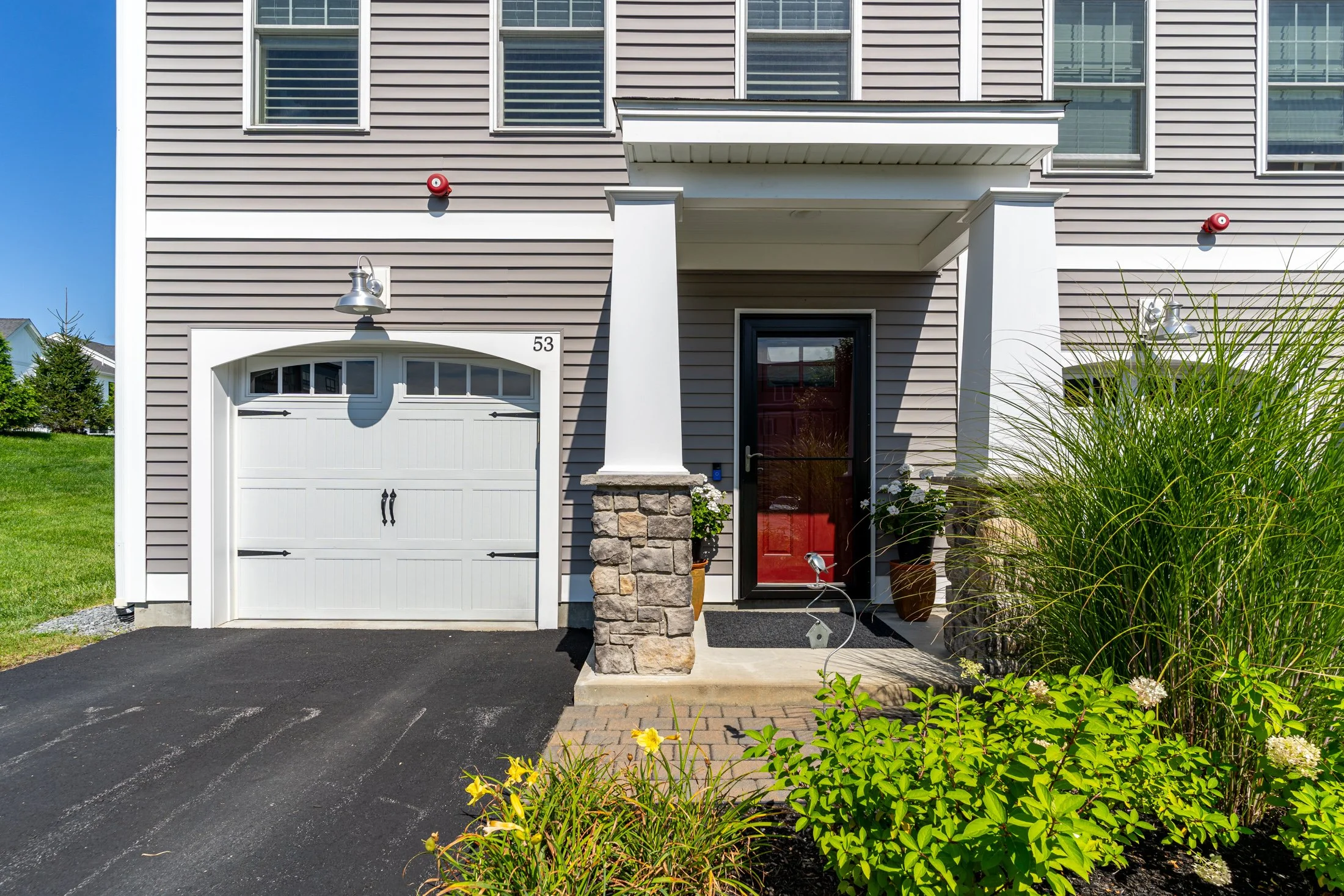 Front view of a modern two-story house with gray horizontal siding, white trim, a red front door, and a white garage door. There are two red security alarm sensors on the house's exterior wall. The house has a small covered porch with a stone pillar, a black mailbox, and outdoor lighting. Green plants and flowers are in front of the porch, and a black asphalt driveway leads up to the garage.