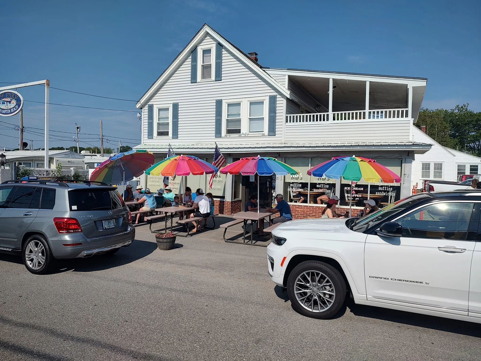 Outdoor dining area with people seated at picnic tables under colorful umbrellas outside a white building with blue shutters, cars parked in front, and a blue sky.