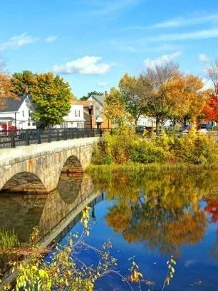 A stone bridge over a river with trees showing fall colors and houses in the background under a partly cloudy sky.