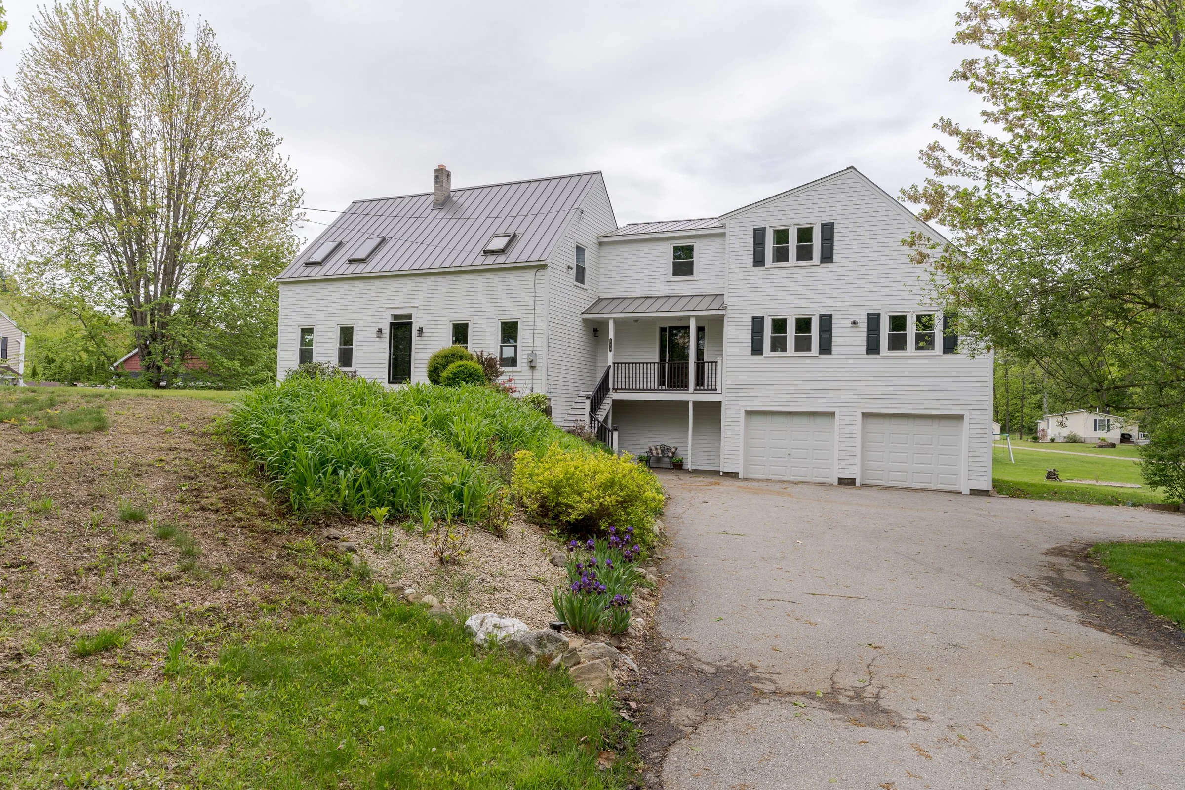 A white two-story house with a metal roof, black window shutters, and a front porch. There is a driveway and a garden with green plants and purple flowers in the front yard, surrounded by trees under a cloudy sky.