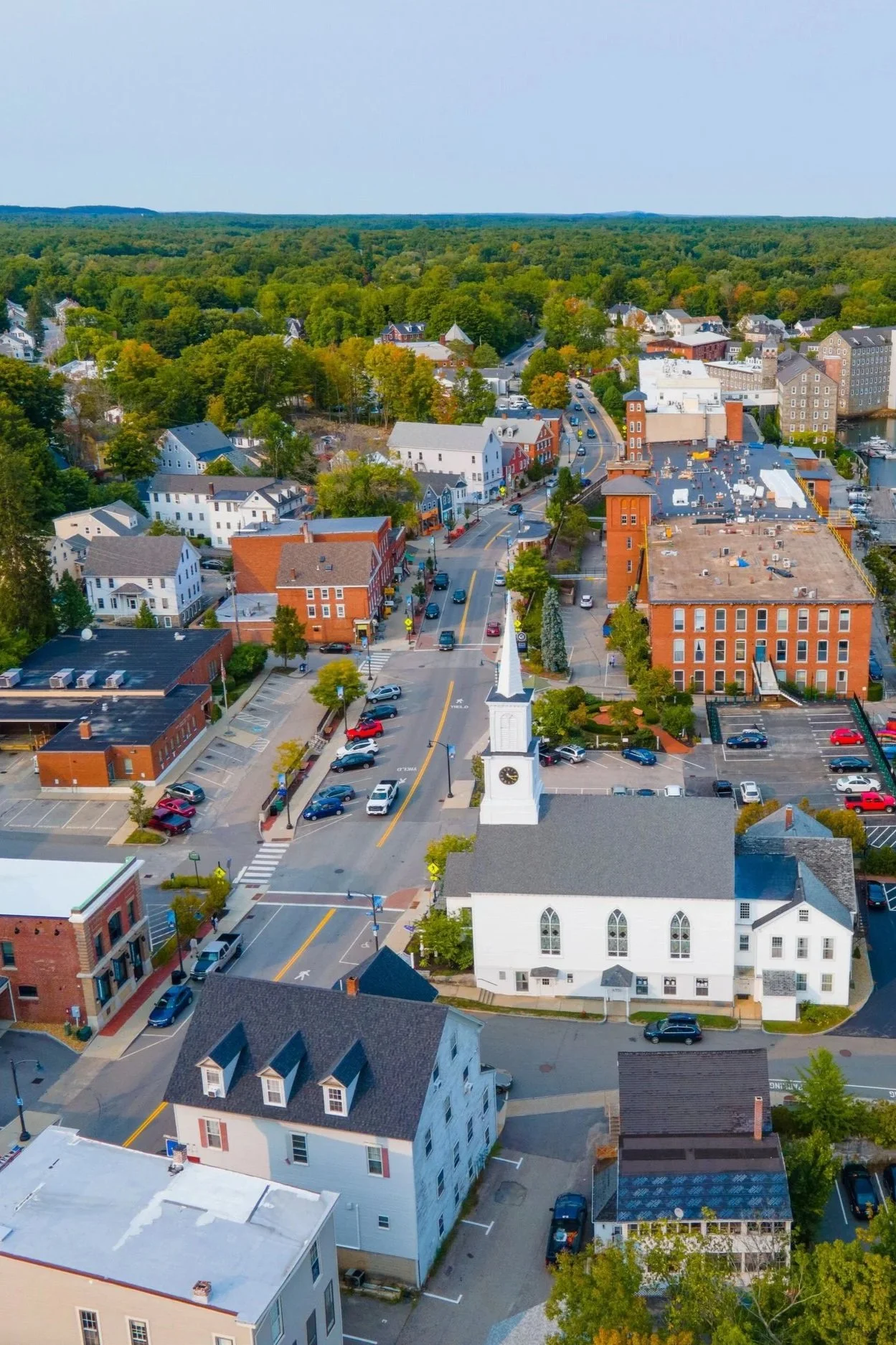 Aerial view of a small town with a white church with a tall steeple, red brick buildings, residential houses, parking lots, and a tree-lined background with a forest and blue sky.