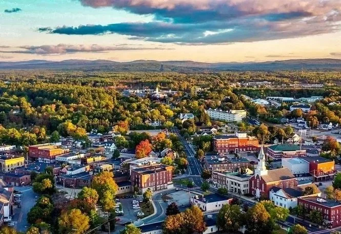 Aerial view of a small town surrounded by trees and rolling hills at sunset, with a mix of historic and modern buildings, streets, and a church steeple.