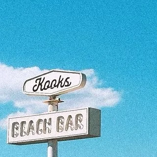 A beach bar sign with the word 'Kooks' above a sign reading 'Beach Bar' against a blue sky with some clouds.