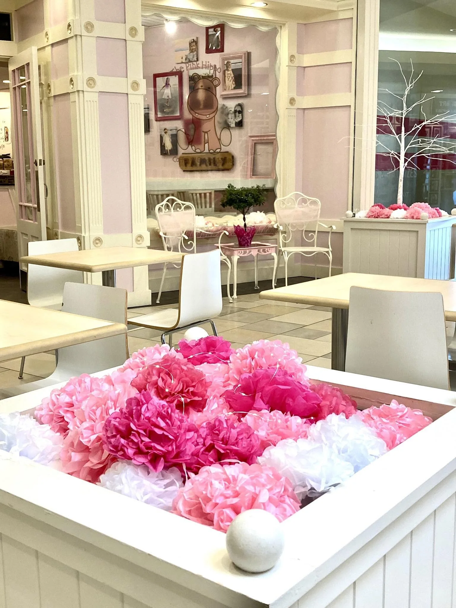 A table decorated with pink and white paper flowers in the foreground. The background shows a cafe or restaurant interior with pastel pink and cream walls, white chairs, and wall art including framed pictures and a cute bear illustration.