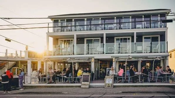 Outdoor dining scene at a multi-story restaurant or cafe during sunset with people seated at tables outside and on upper balcony, in an urban setting.